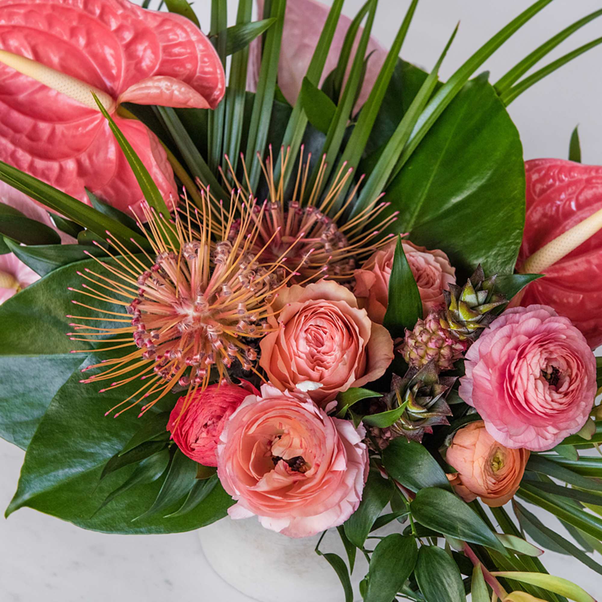 Tropical arrangement with pink anthuriums, roses, and protea in a white vase