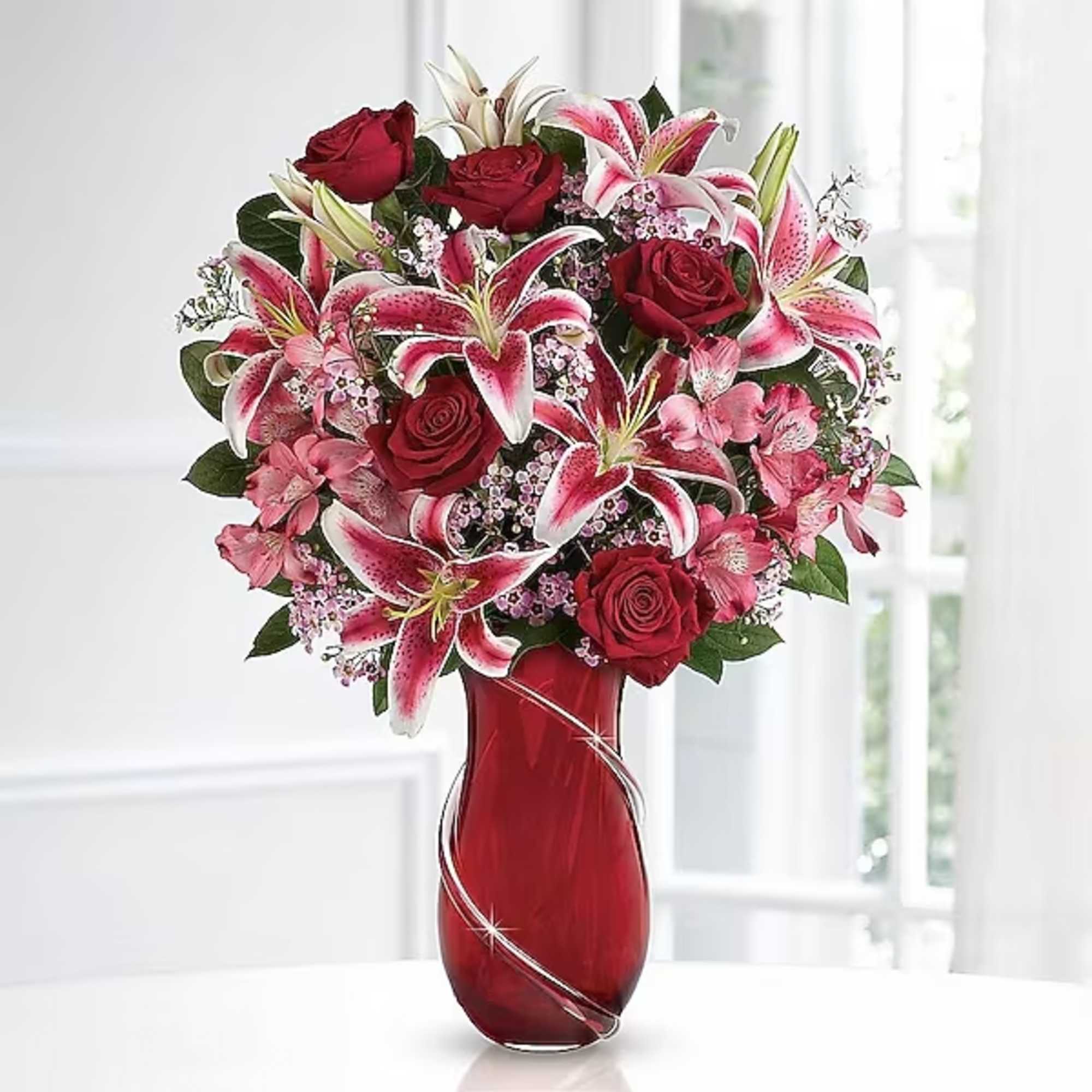 Red and pink lilies and roses arranged in a red glass vase with silver swirl accents