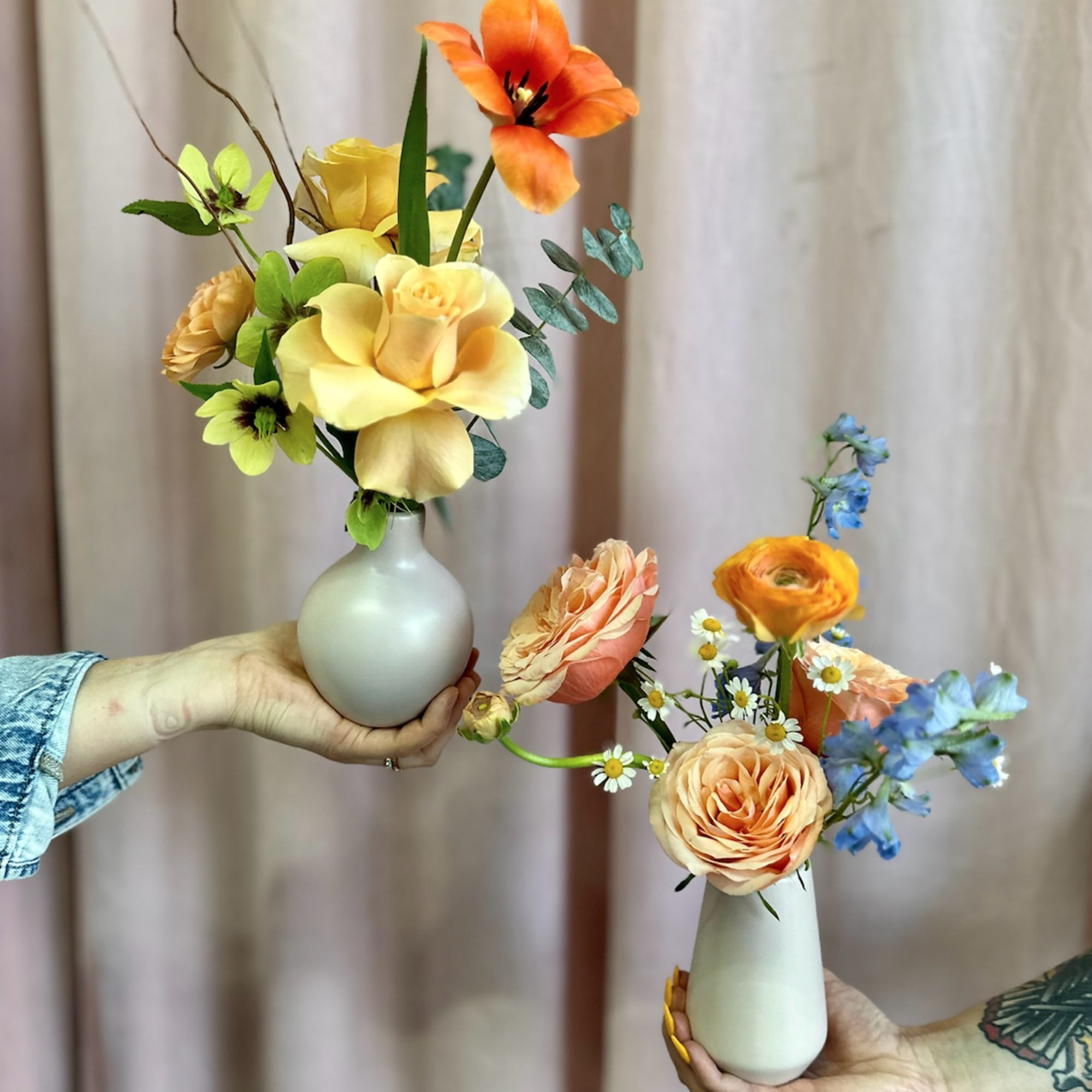Two small pastel vases with yellow, peach, and blue flowers held by hands against a pink backdrop