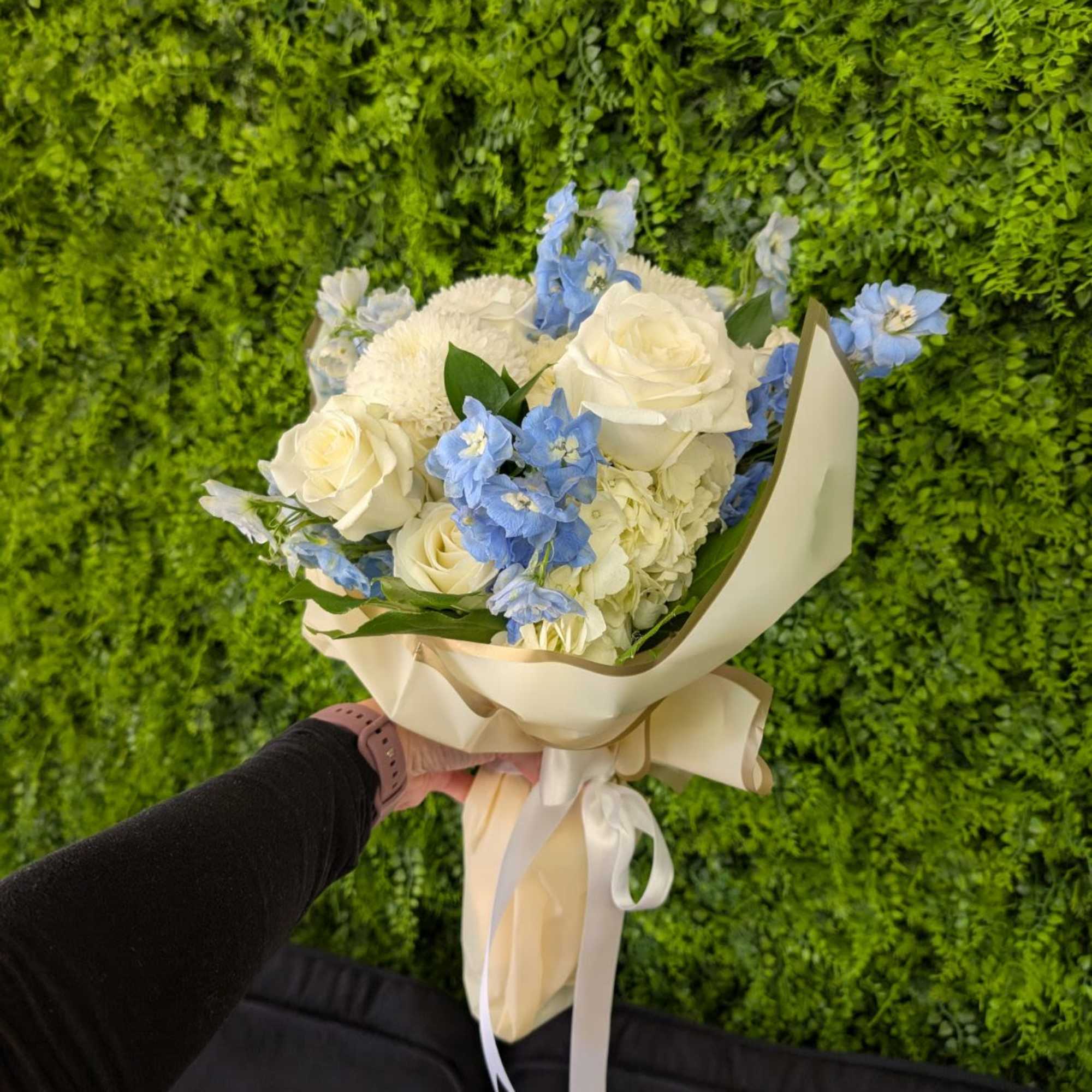 Hand-tied bouquet of white roses and blue flowers in cream wrap with ribbon, held against a green wall.