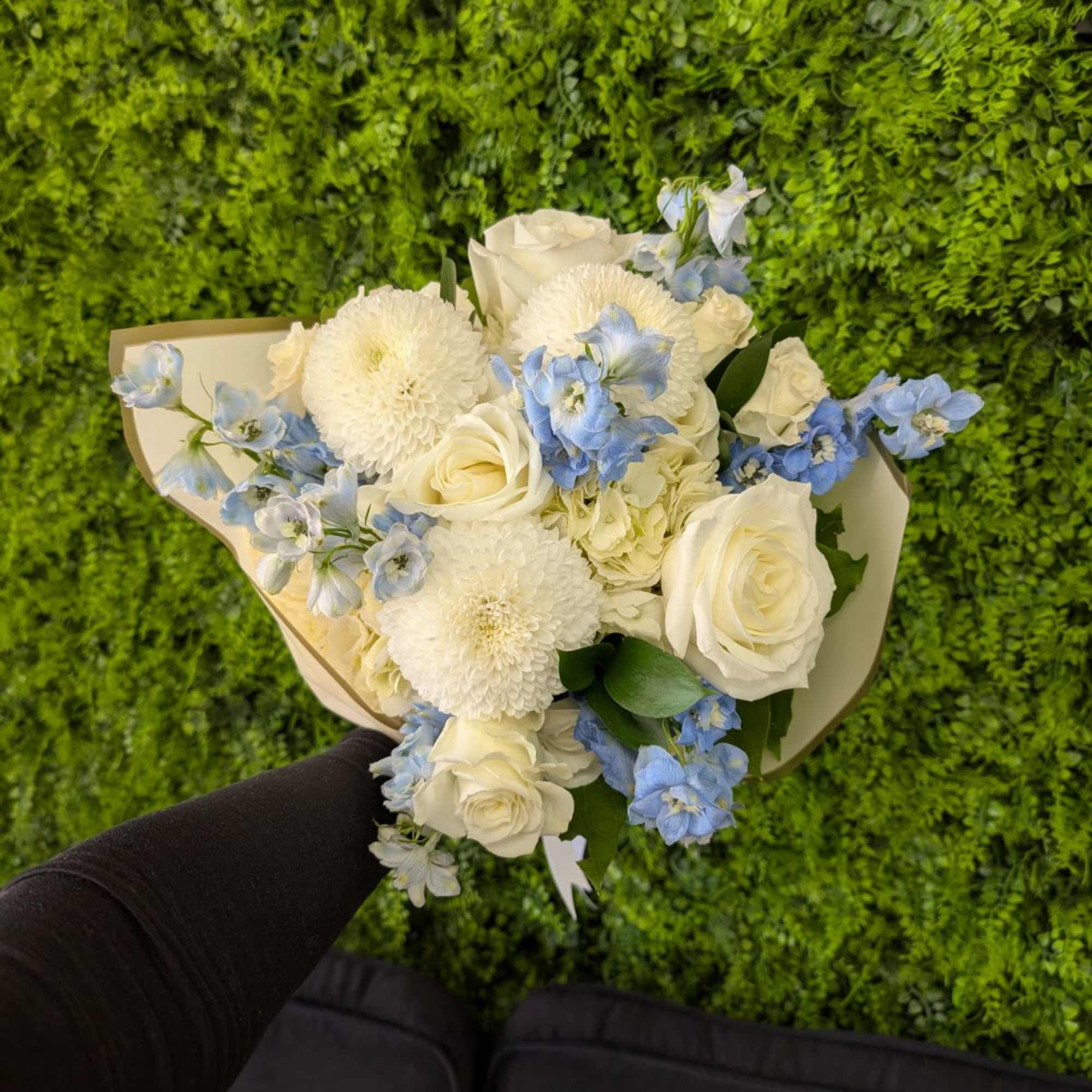 Handheld bouquet of white roses and round blooms with pale blue flowers wrapped in cream paper.