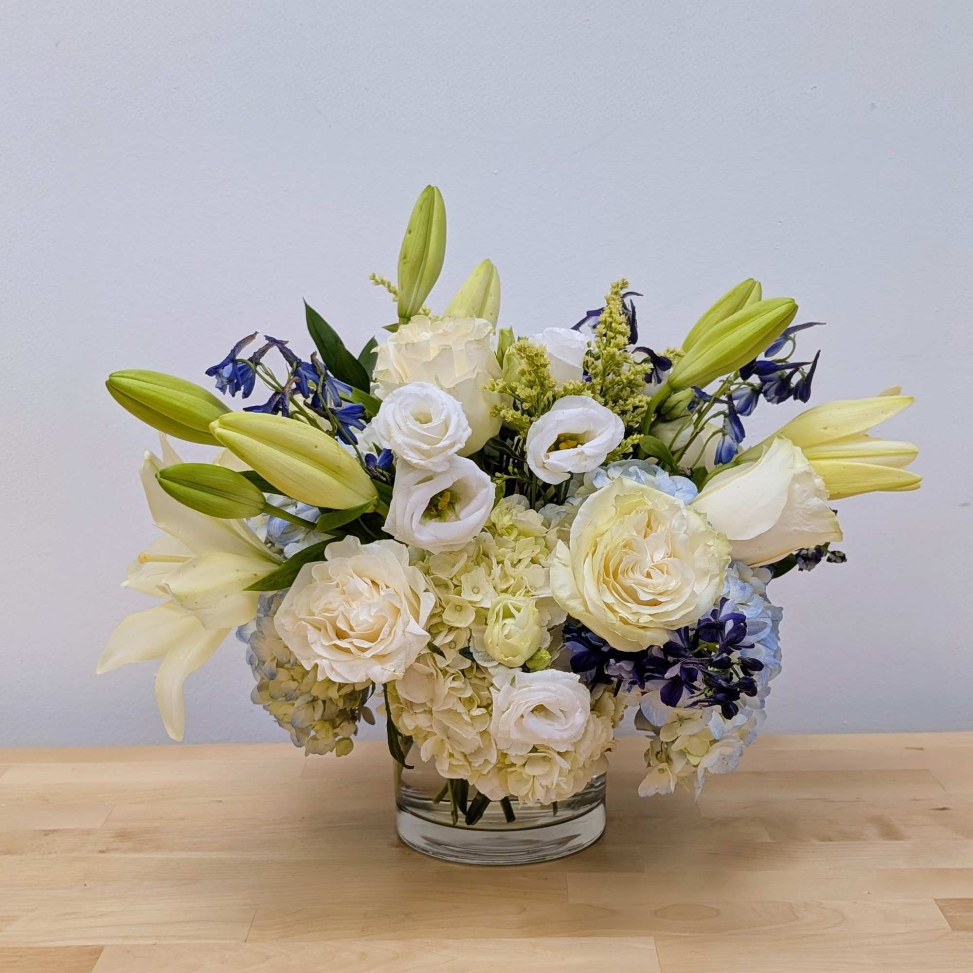 Low glass vase of white roses, lilies, hydrangeas and blue accent flowers in a rounded arrangement