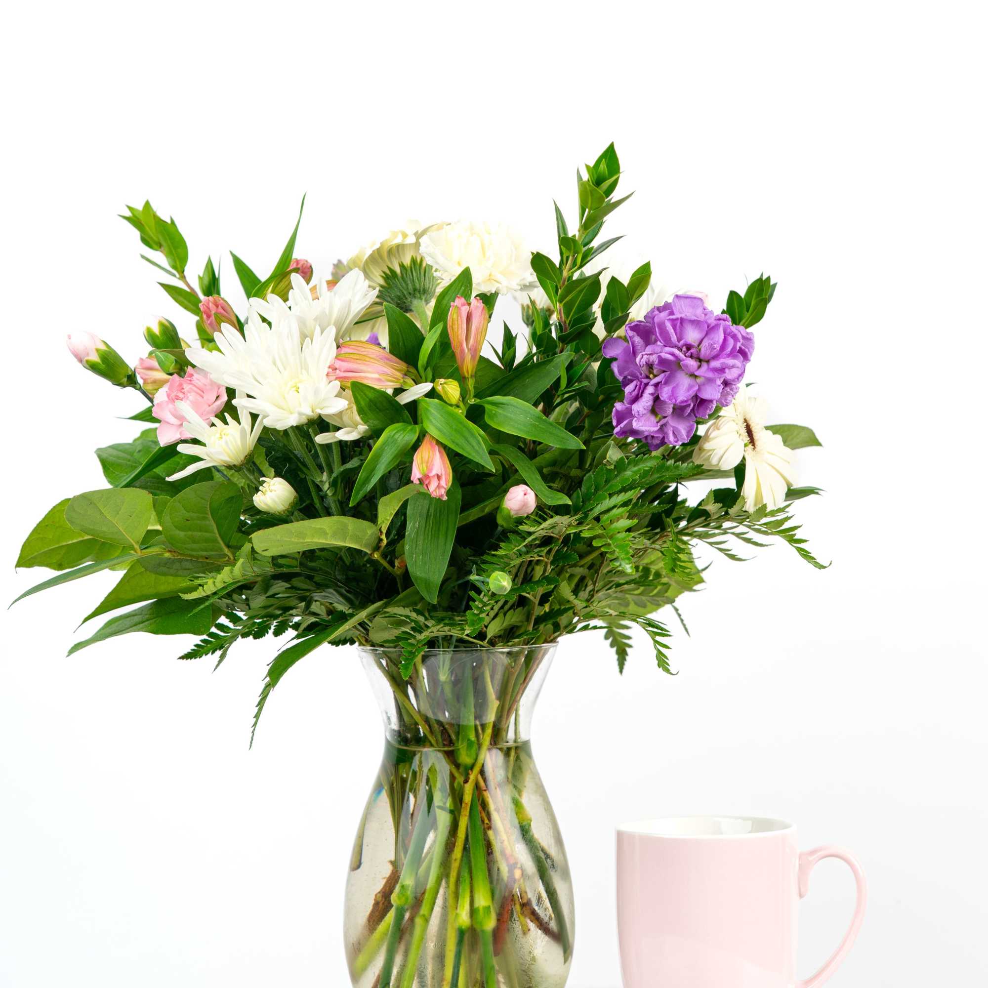 Mixed bouquet of white, pink, and purple flowers in a clear vase beside a pink mug