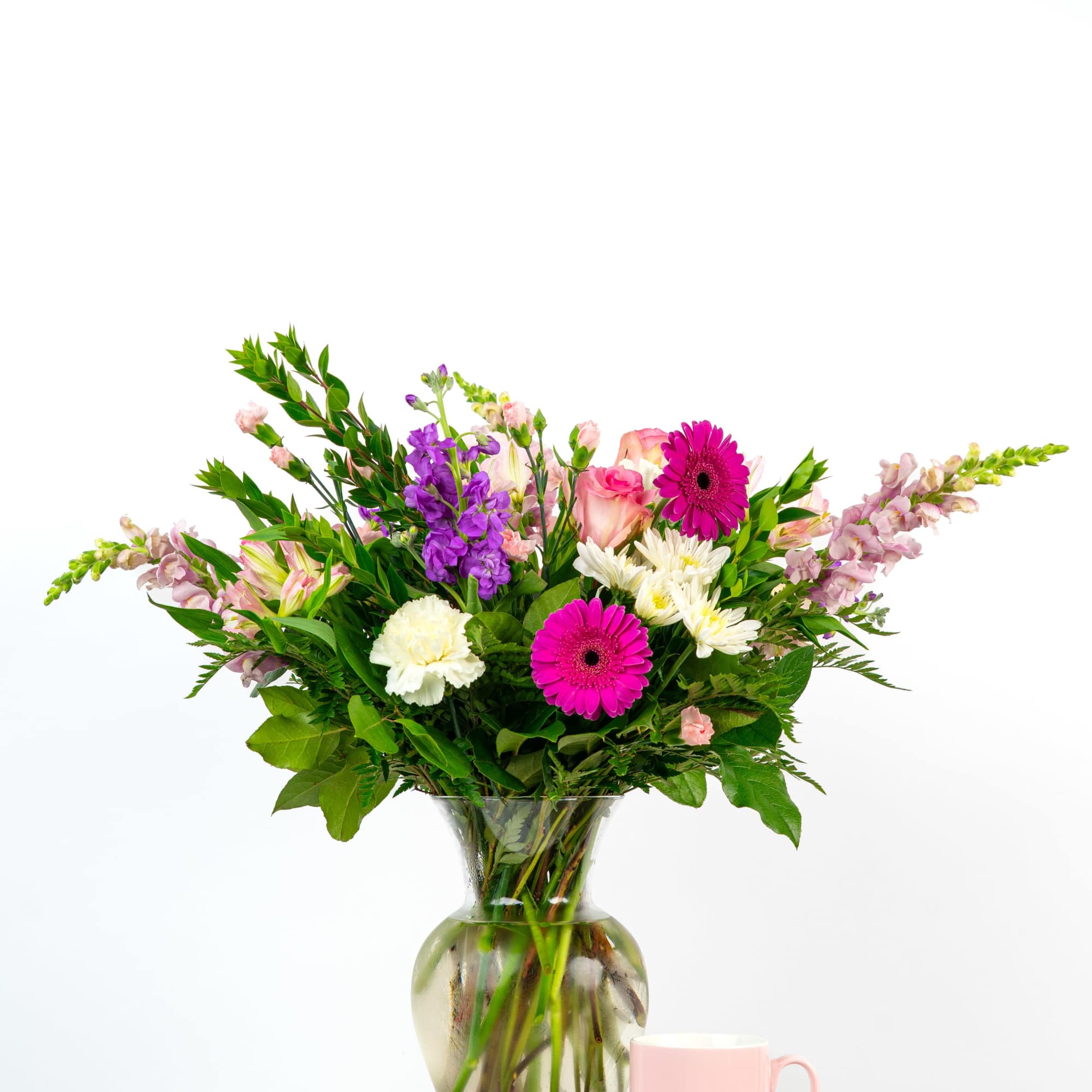 Tall mixed bouquet of pink, purple, and white flowers in a clear vase beside a pink mug