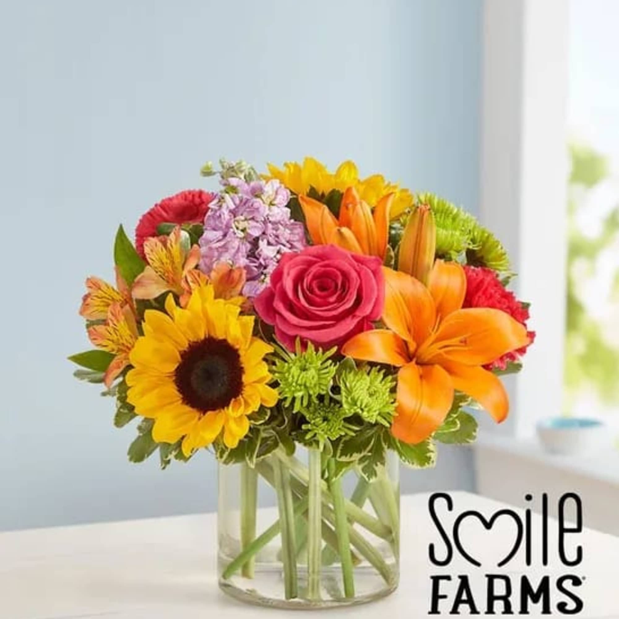 Bright mixed bouquet with sunflowers, orange lilies, pink rose and other blooms in a clear glass vase.