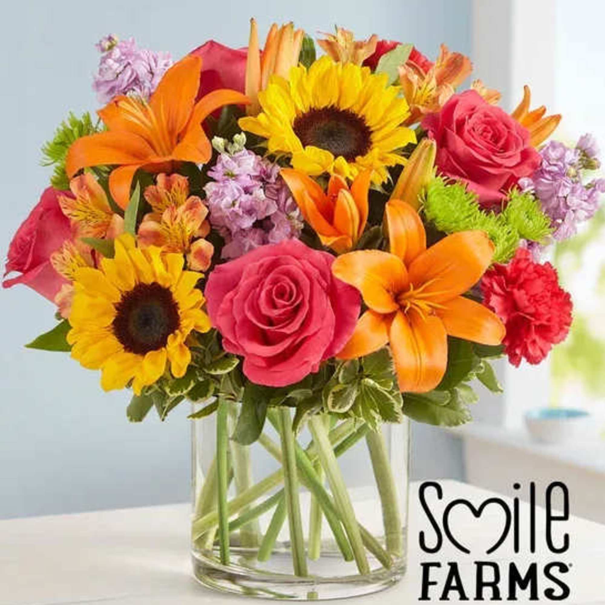 Bright mixed bouquet with pink roses, yellow sunflowers, and orange lilies in a clear glass cylinder vase