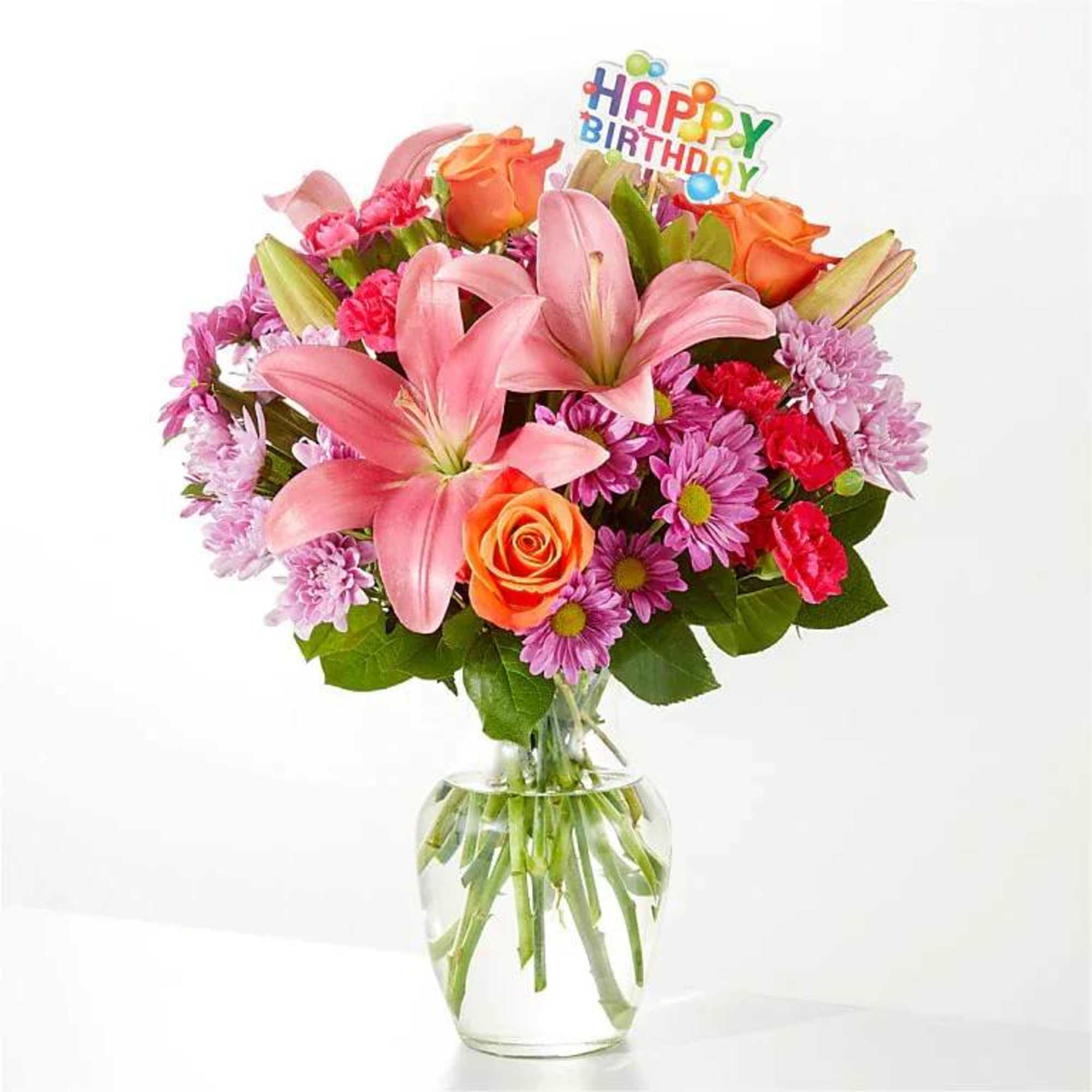 Mixed bouquet of pink lilies, orange roses, and purple mums in a clear glass vase with a Happy Birthday pick.