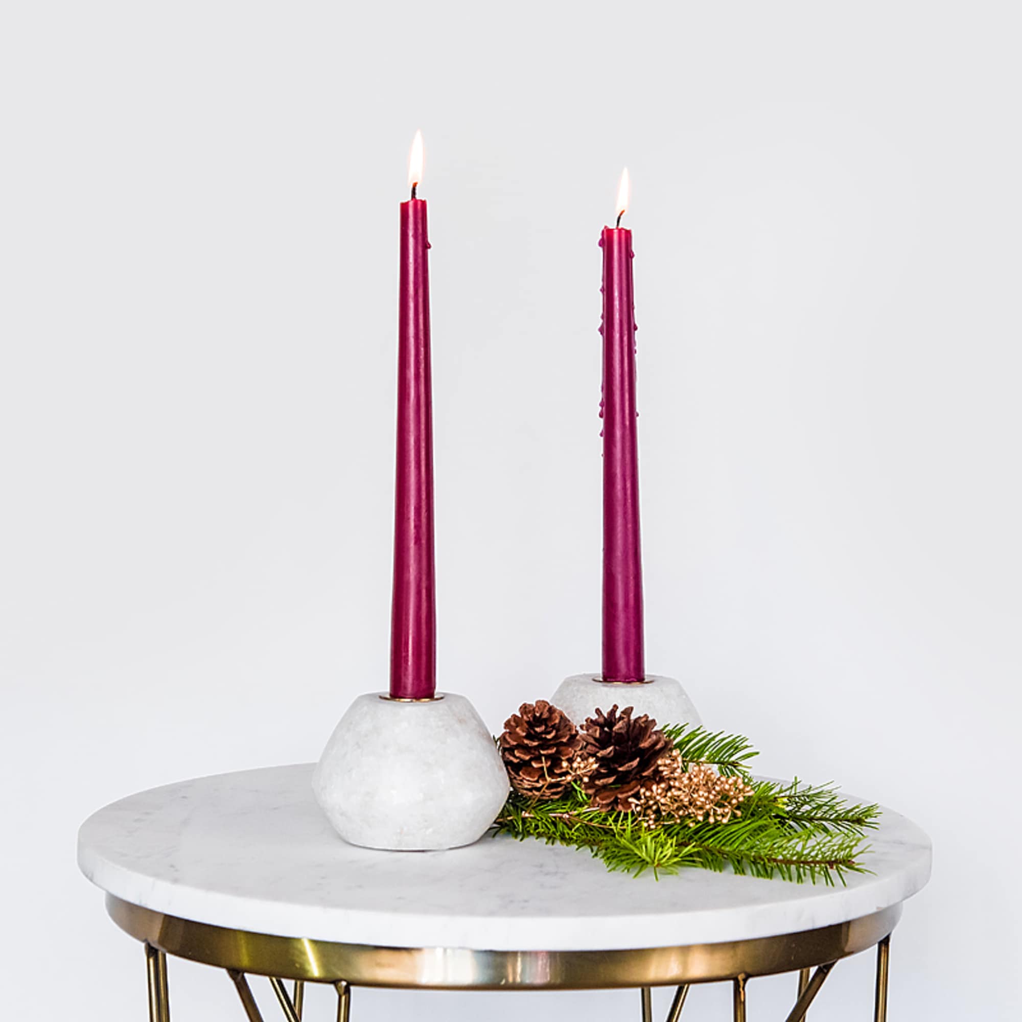 Two burgundy taper candles in white holders with pinecones and evergreens on a marble table