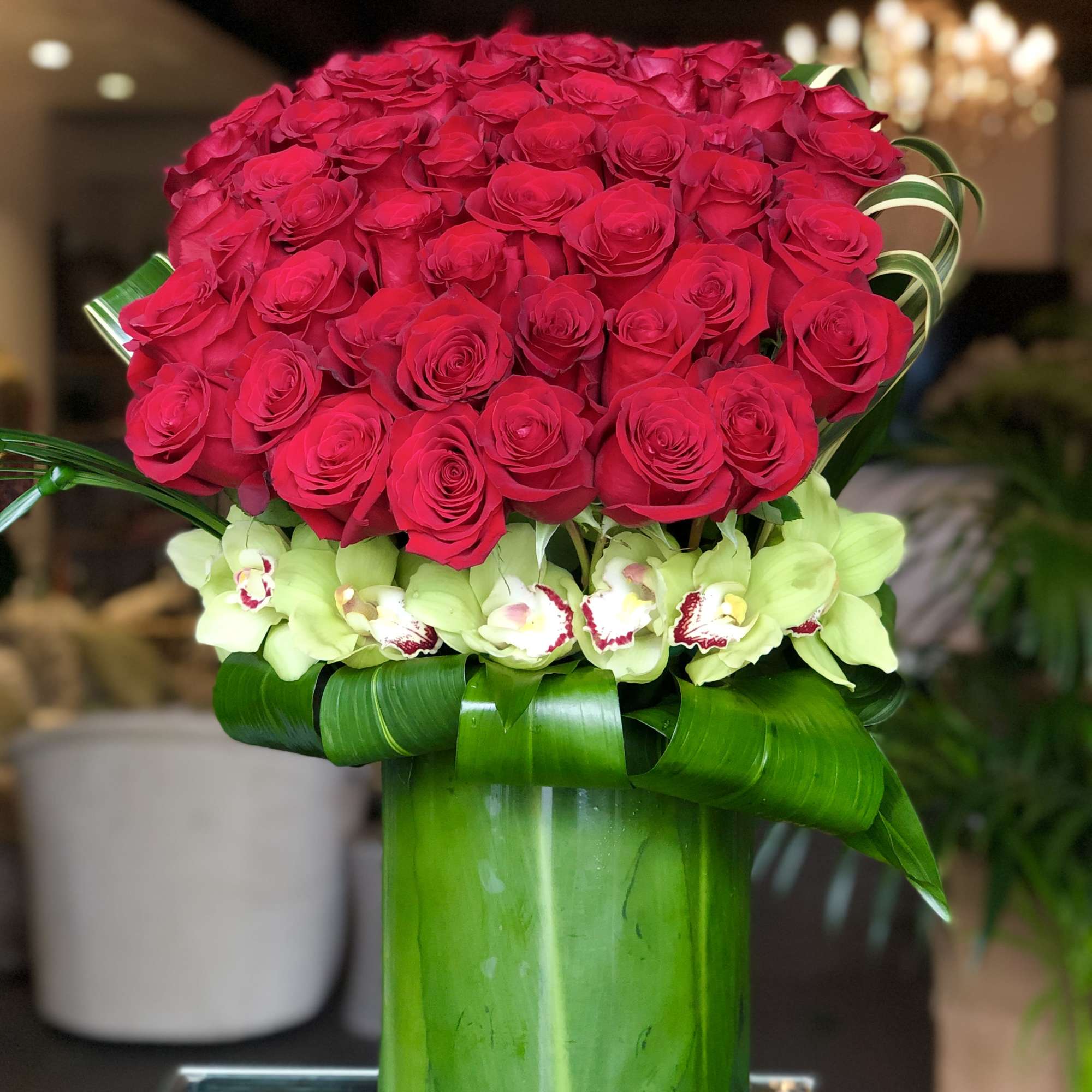 Tall arrangement of red roses with green orchids in a leaf-wrapped glass cylinder vase