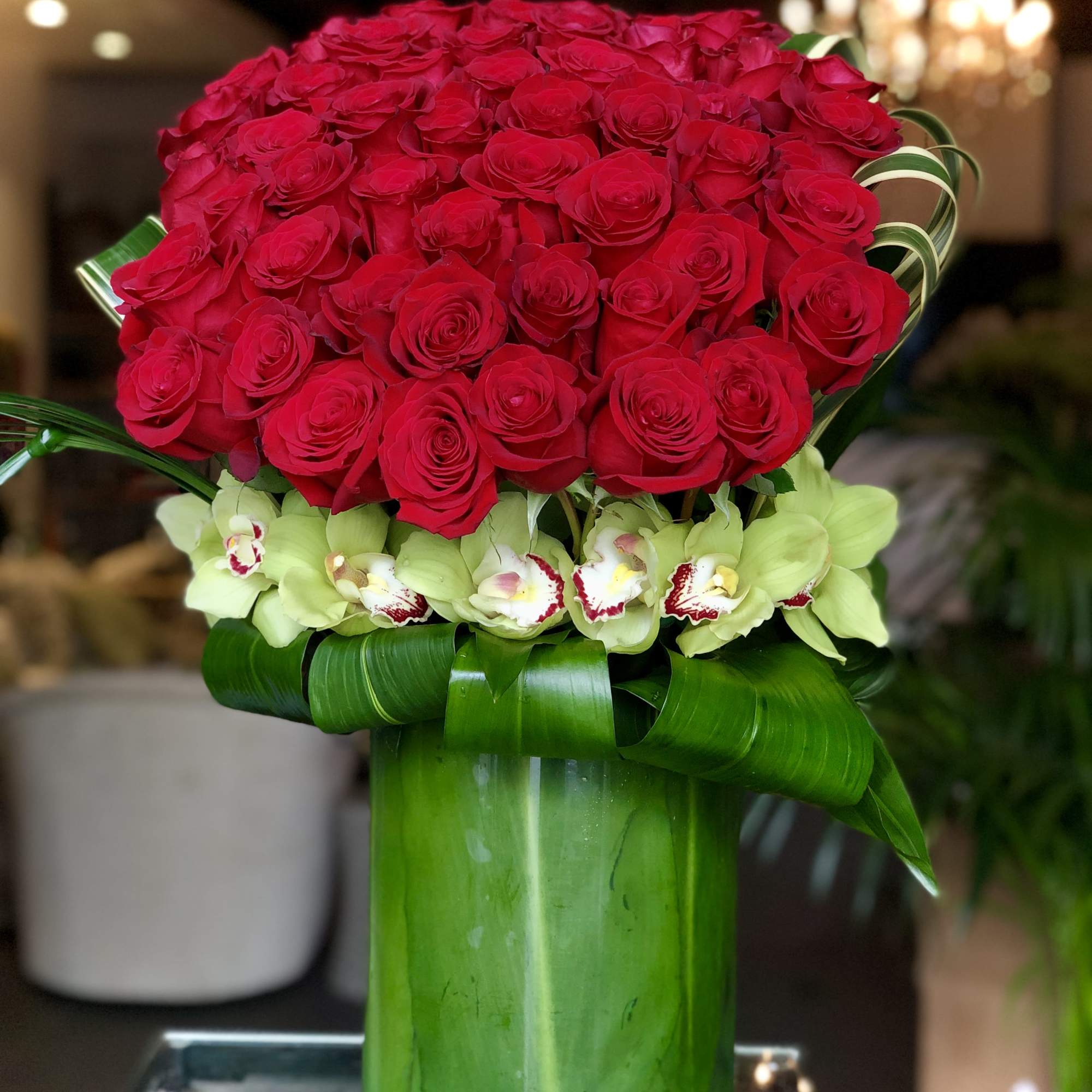 Tall arrangement of red roses with green orchids in a green-wrapped glass cylinder vase