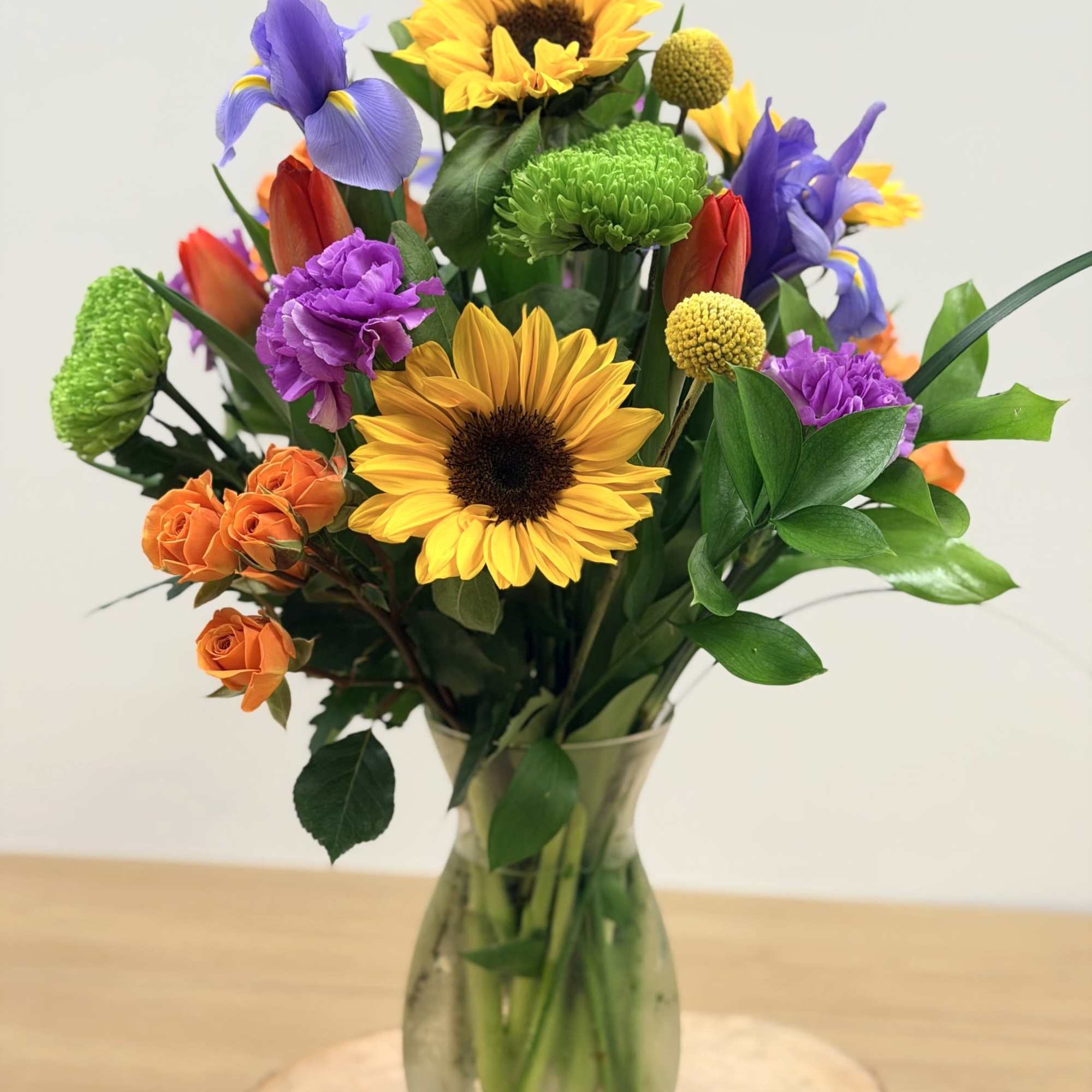 Tall mixed arrangement with sunflowers, iris, orange roses, tulips and green mums in a clear glass vase
