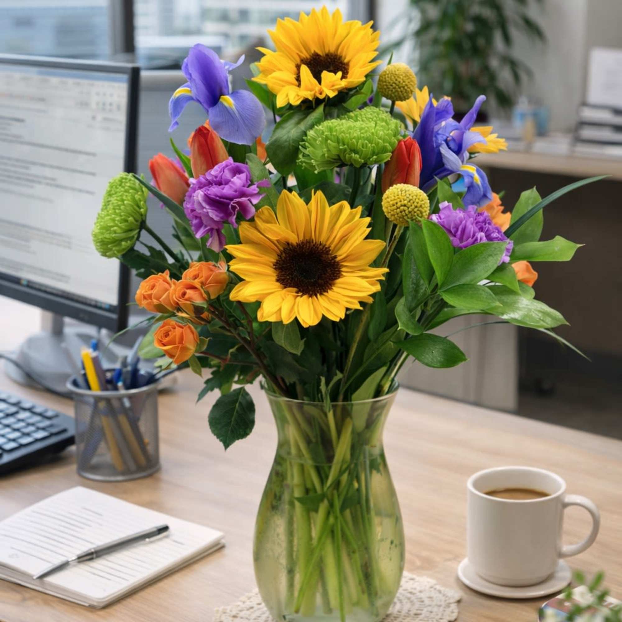 Mixed bouquet with sunflowers, purple irises, tulips, and roses in a clear glass vase on an office desk
