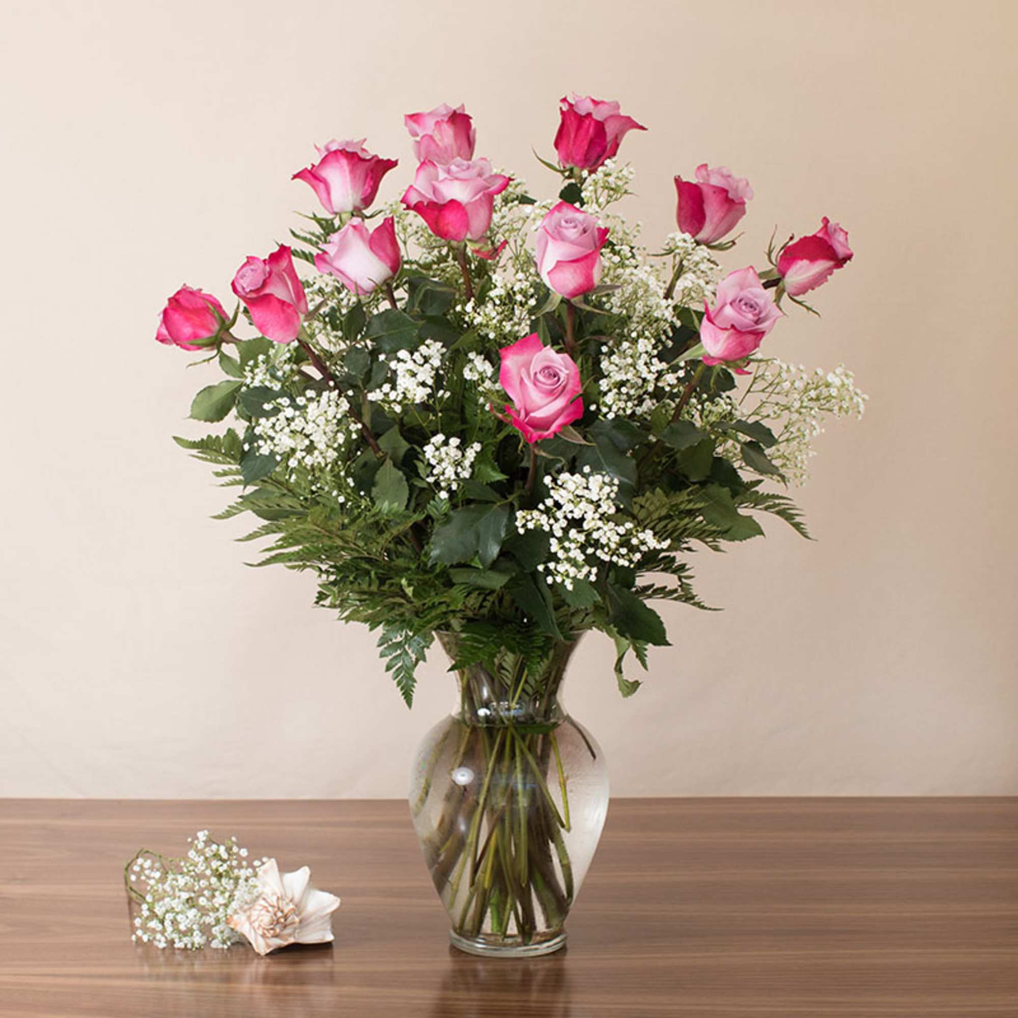 Tall arrangement of pink roses with white filler flowers in a clear glass vase on a wooden table