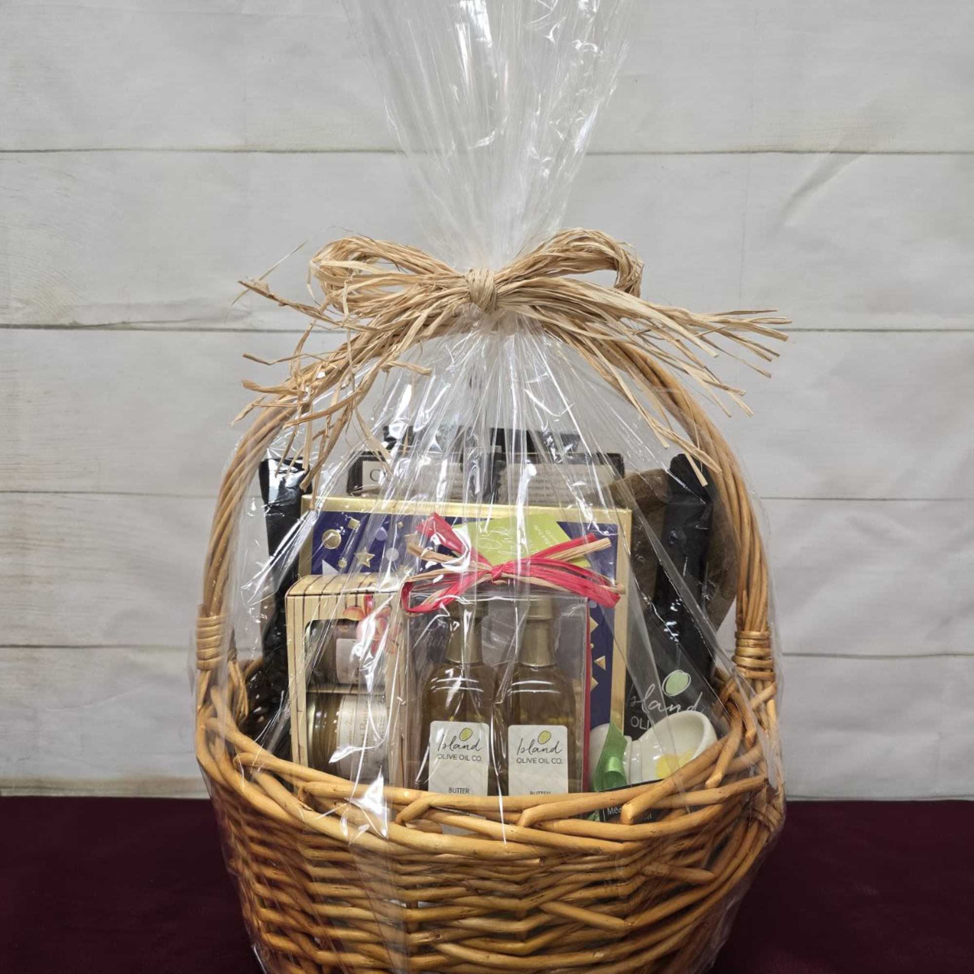 Large wicker gift basket wrapped in clear cellophane and tied with a raffia bow, filled with assorted gourmet items.