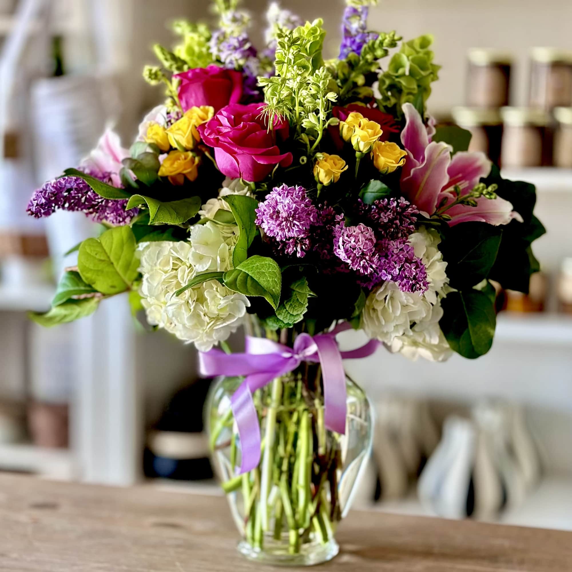 Mixed bouquet of roses, lilies, hydrangeas, and lilacs in a glass vase with a purple ribbon