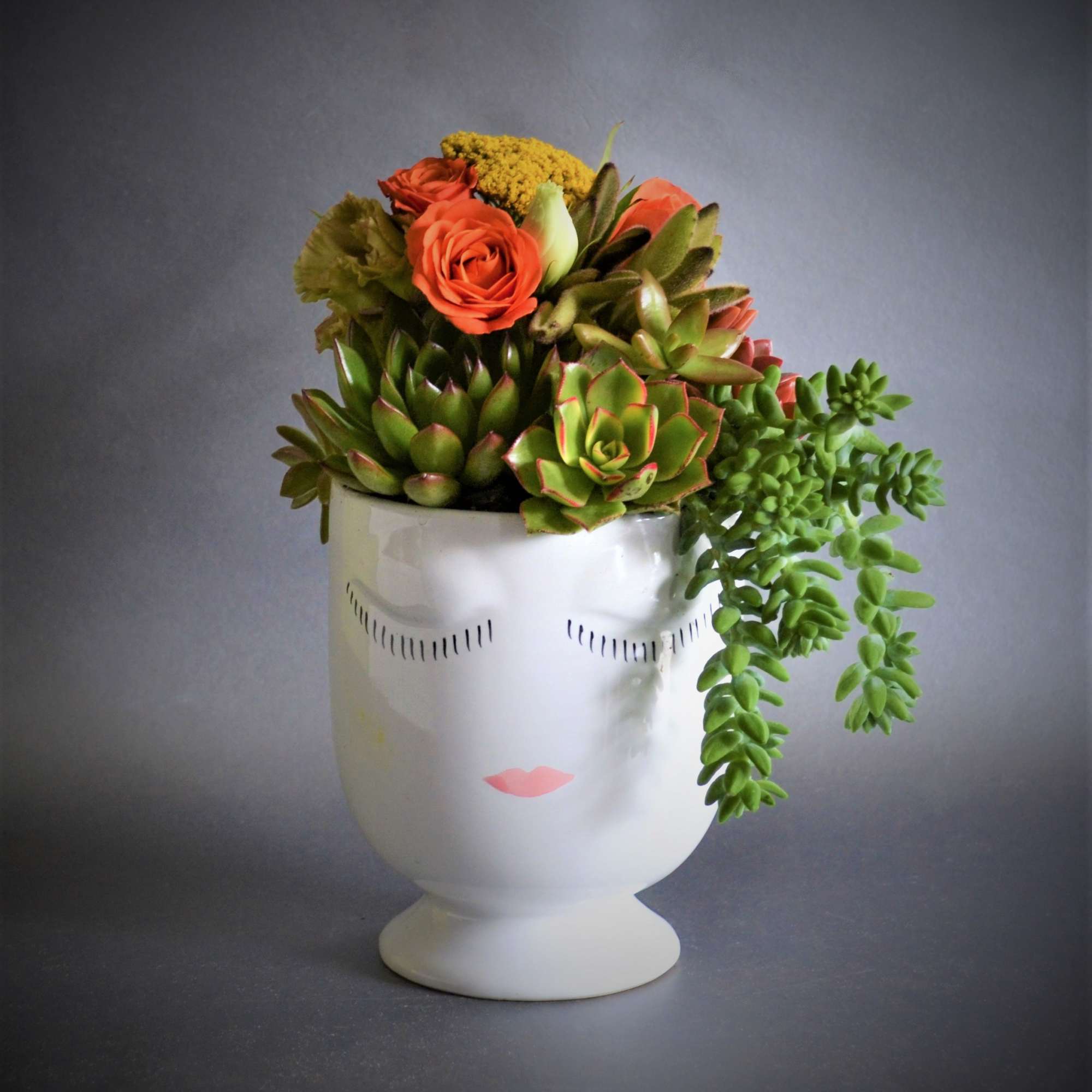 Succulent arrangement with orange roses in a white face vase
