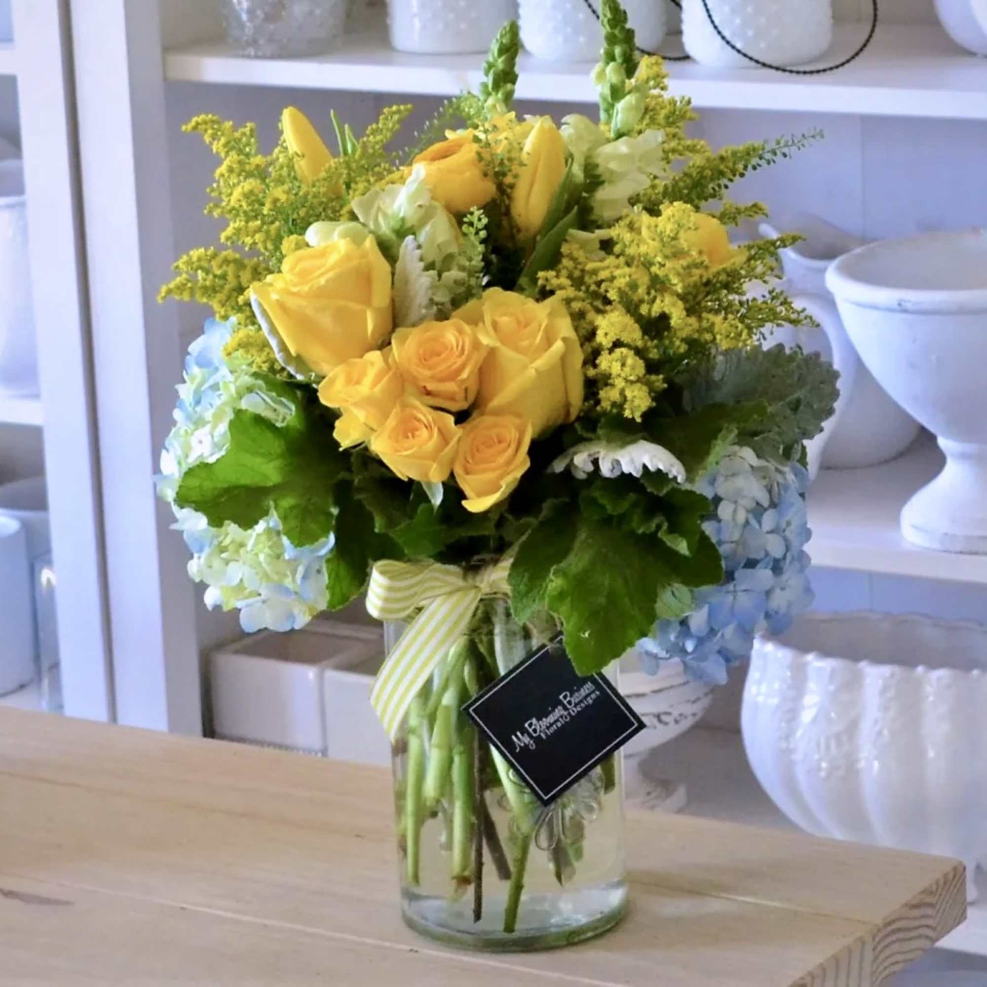 Yellow roses and blue hydrangeas in a glass vase with a striped ribbon.