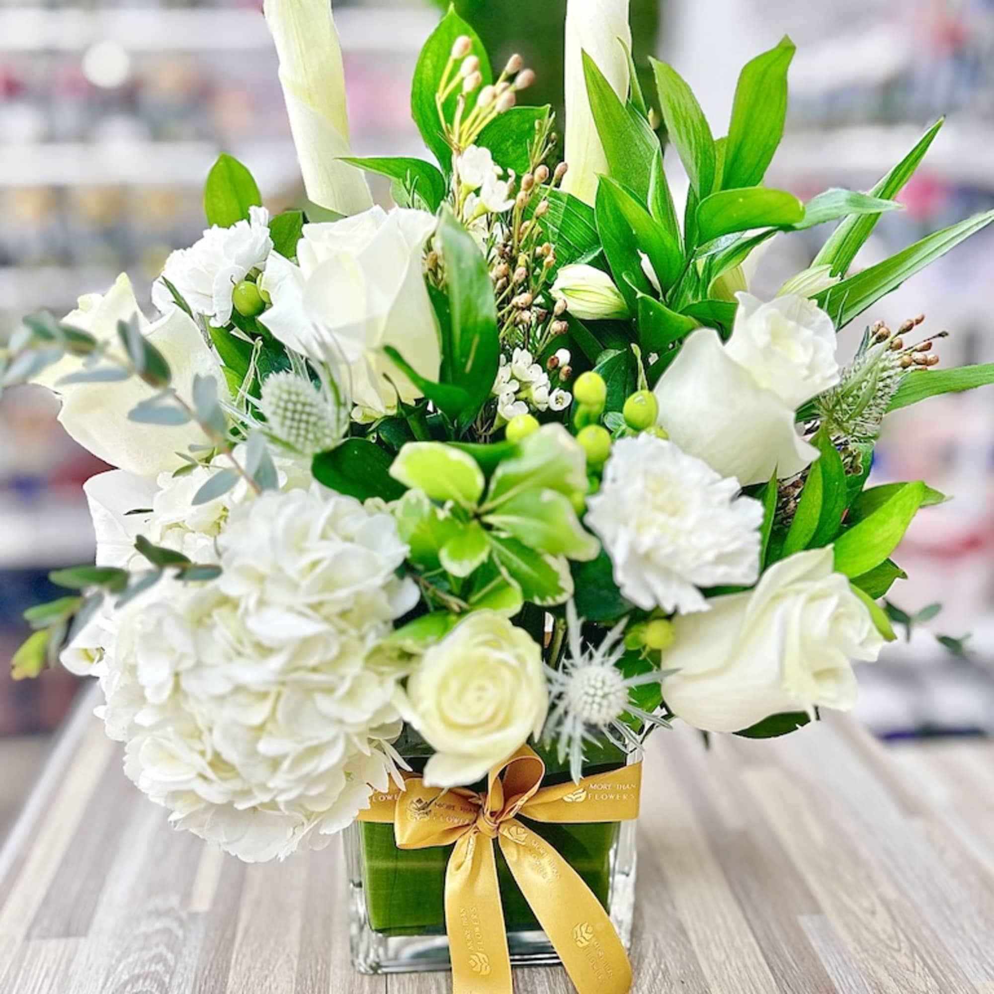 White and green flower arrangement with roses, hydrangea, and calla lilies in a glass cube vase with yellow ribbon