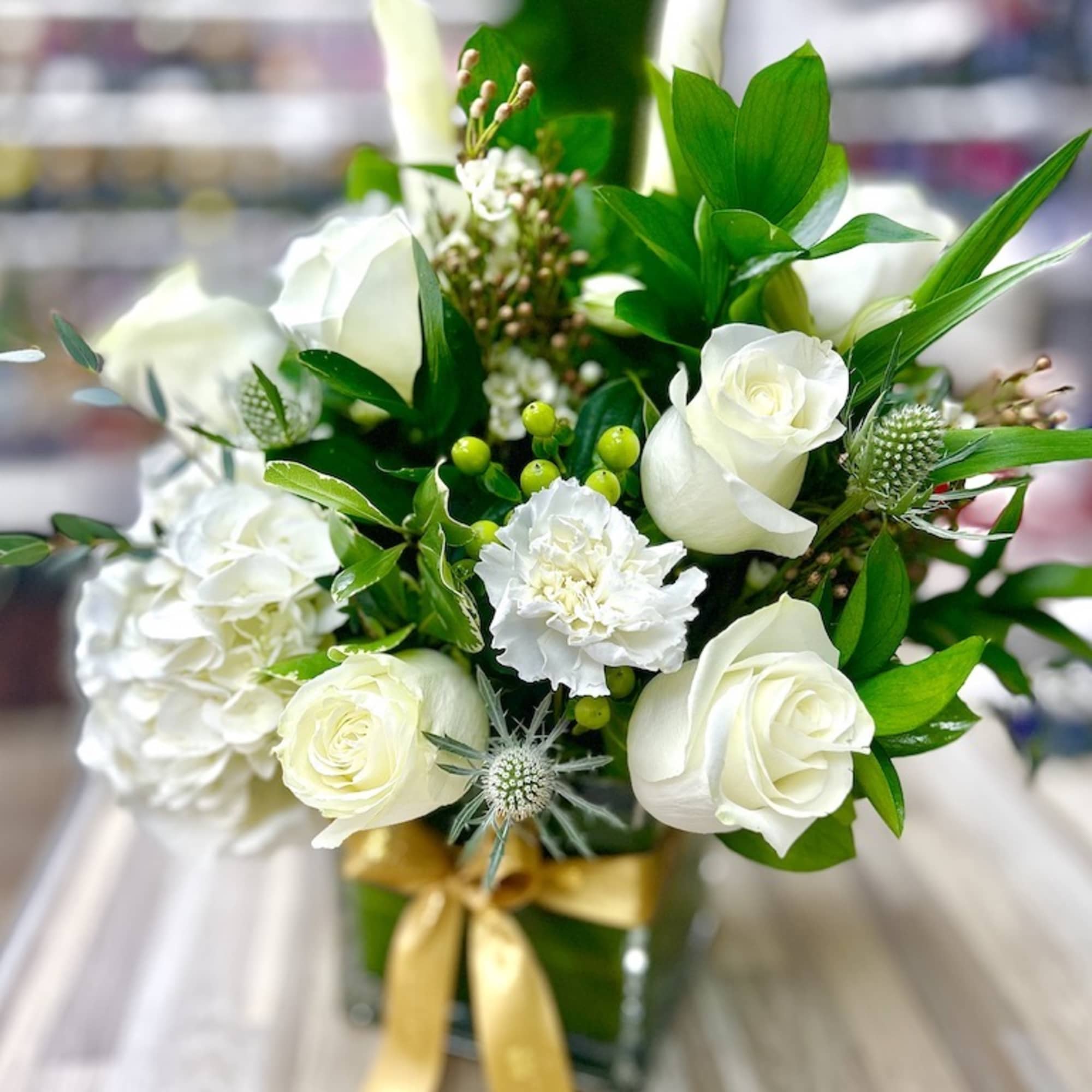 White roses, hydrangea, calla lilies, and carnations in a glass cube vase with a gold ribbon