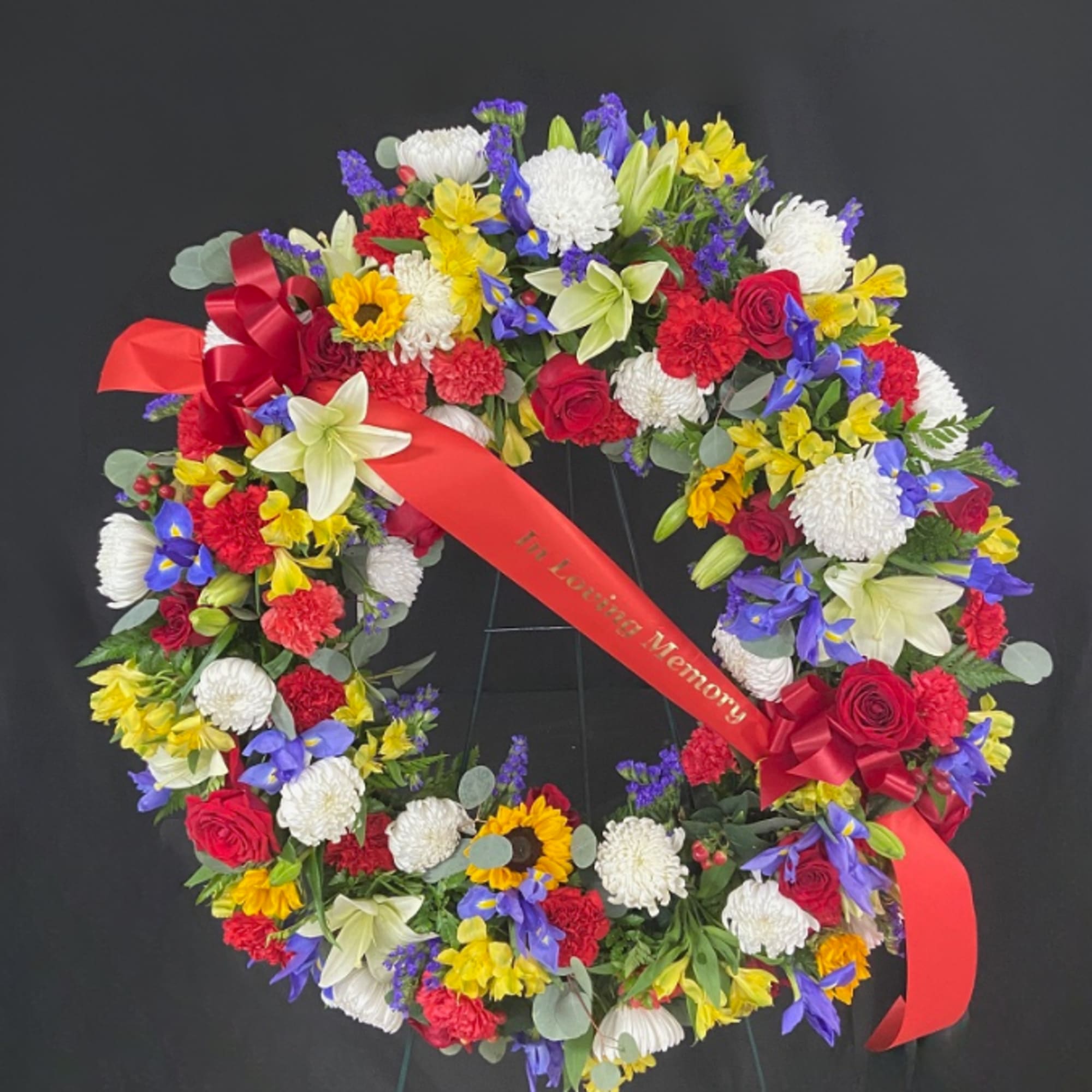 Colorful circular sympathy wreath with red, white, yellow, and blue flowers on a stand with a red memorial ribbon.