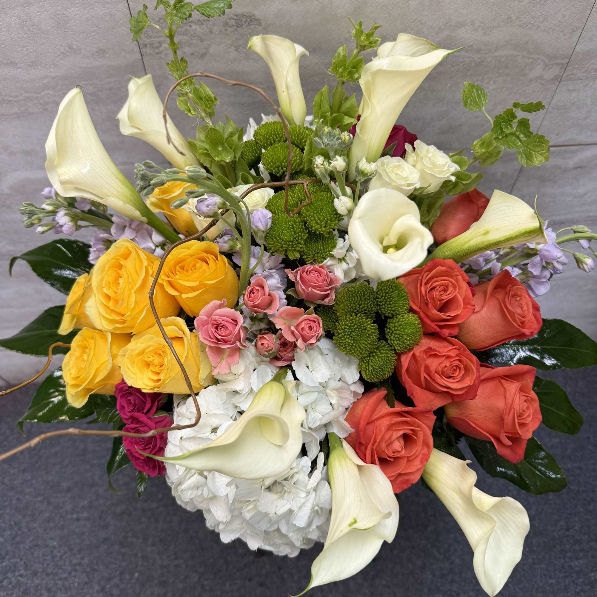 Mixed arrangement of white calla lilies, hydrangeas, and colorful roses in a round white vase