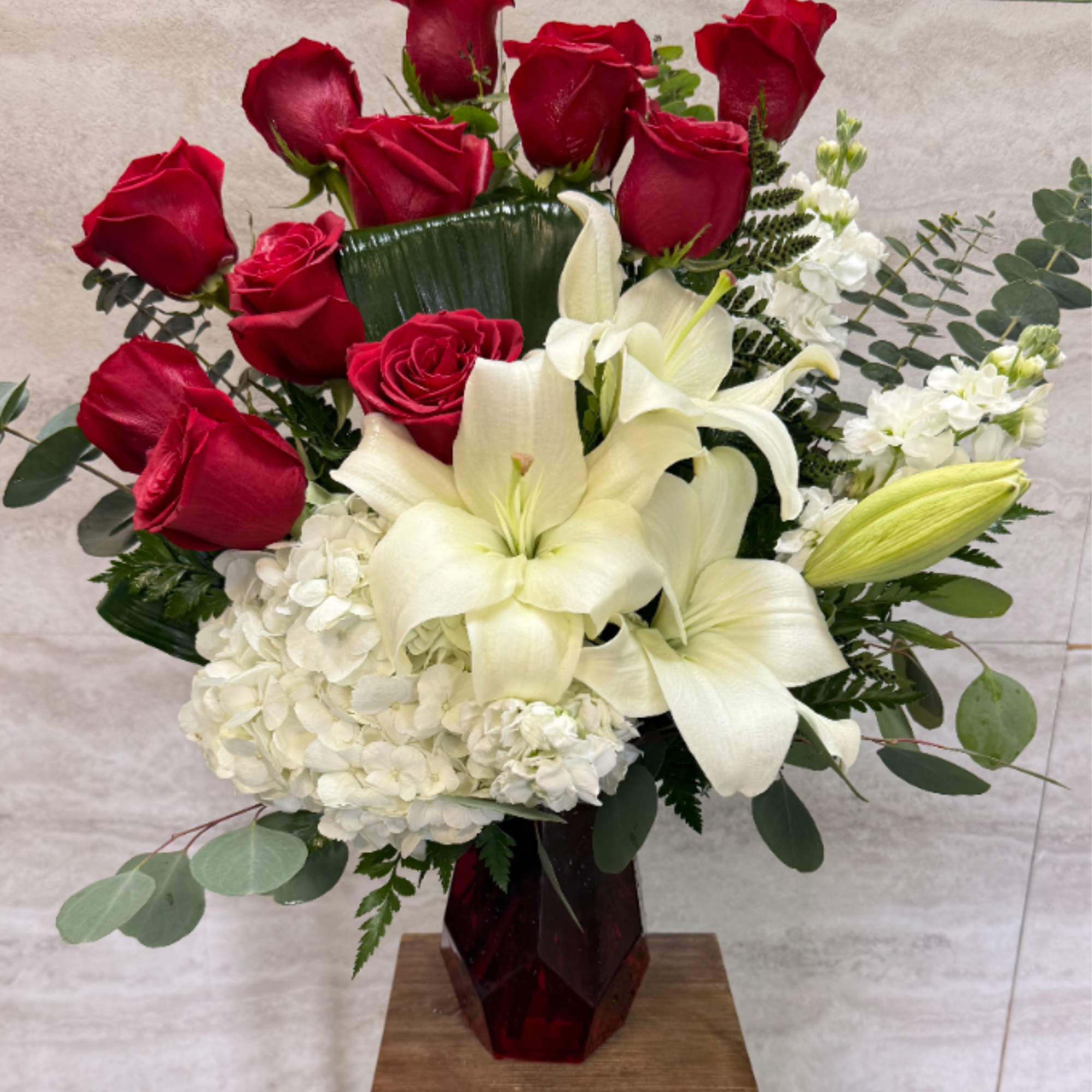 Tall arrangement of red roses, white lilies, and white hydrangeas in a red glass vase