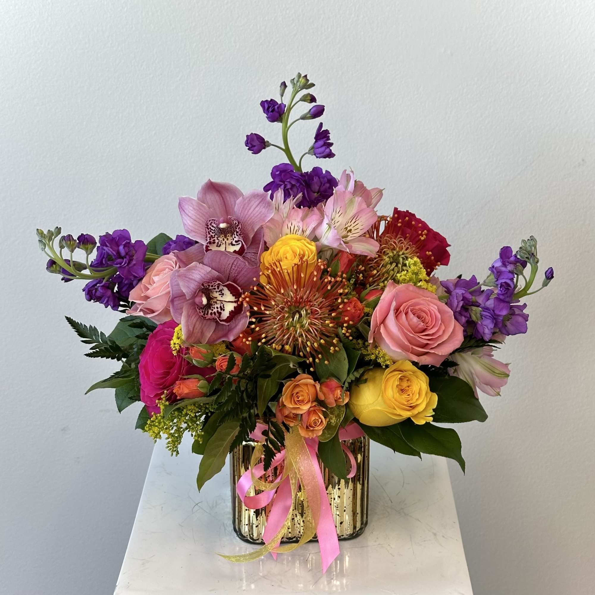 Colorful bouquet of roses, orchids, and mixed blooms in a gold vase with pink ribbon