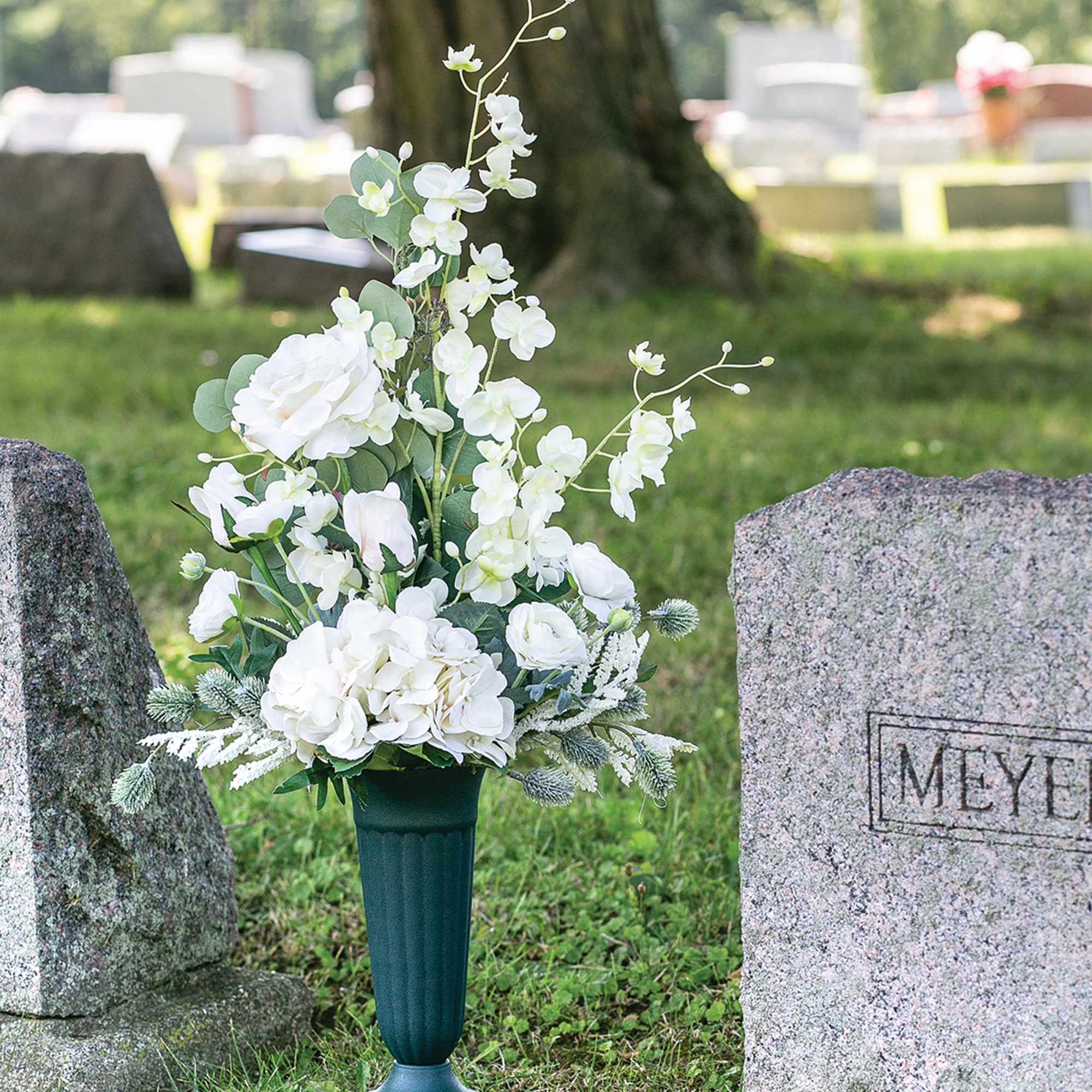 White floral arrangement in a dark green cemetery vase beside gravestones