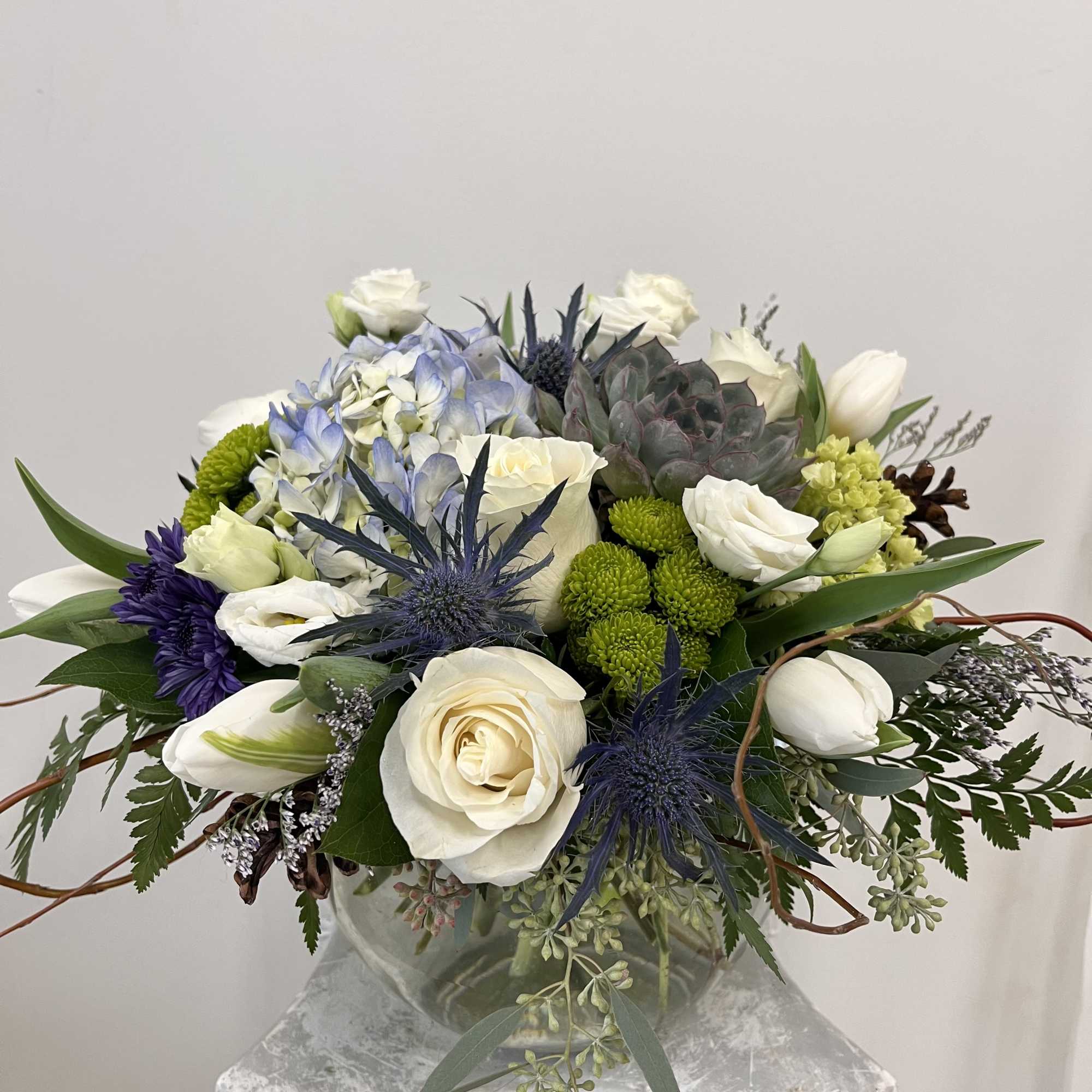 Round arrangement of white roses, tulips, blue hydrangea and succulents in a clear glass vase