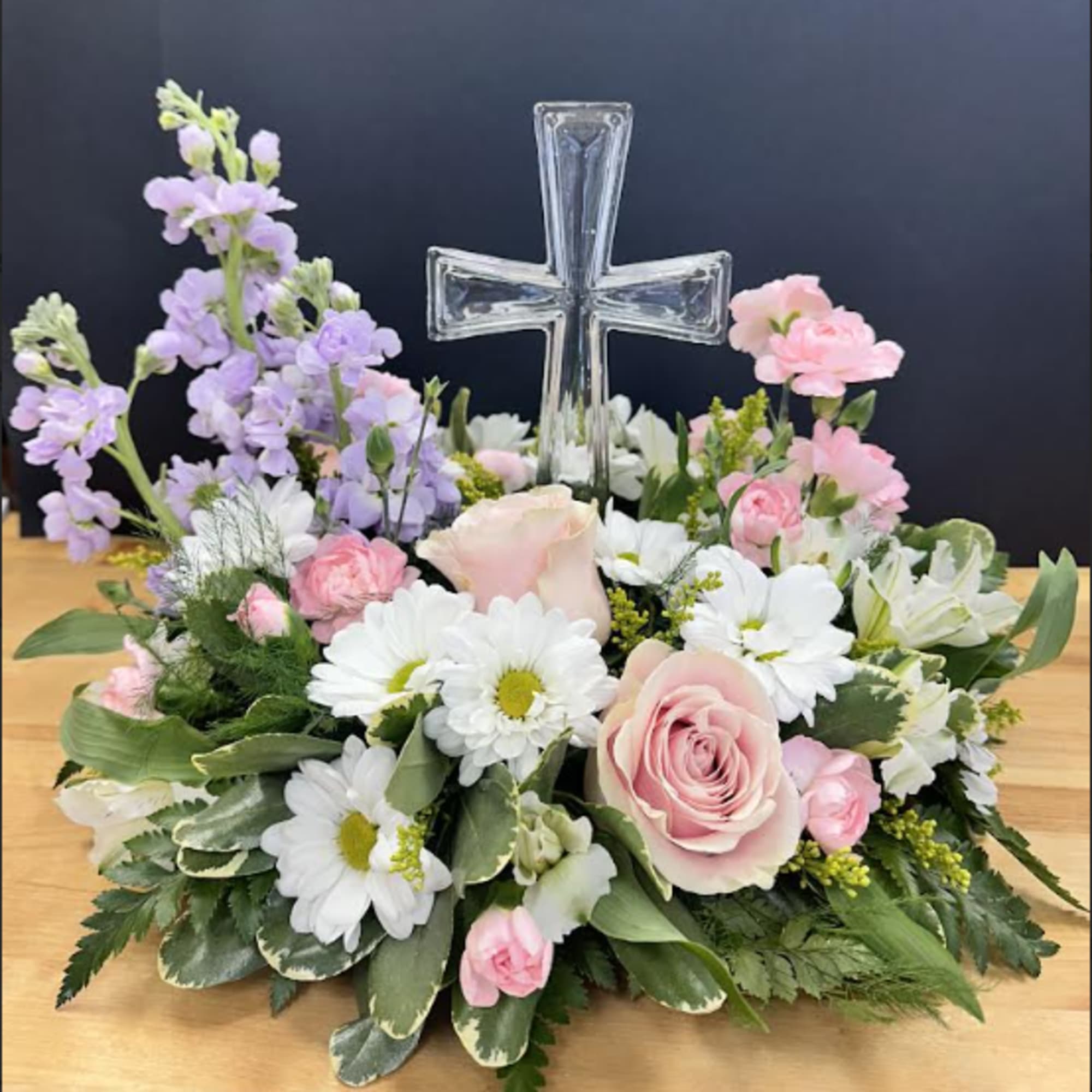 Low arrangement of pink roses, white daisies, and lavender blooms surrounding a clear glass cross.