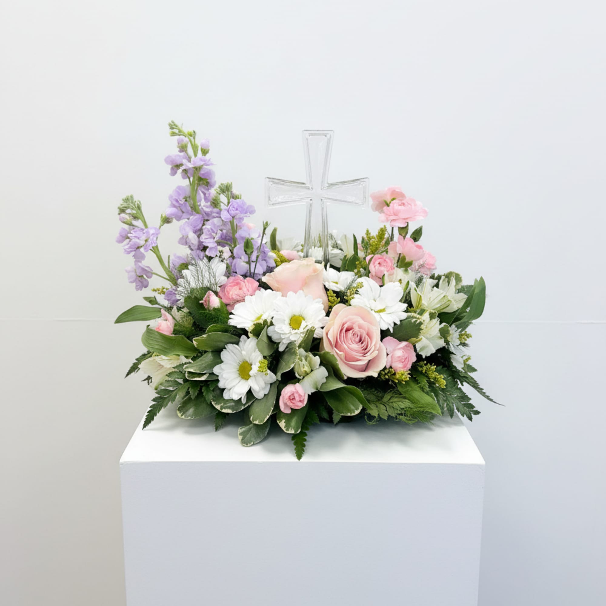 Low pastel arrangement of pink roses, white daisies, and lavender blooms with a clear glass cross in the center