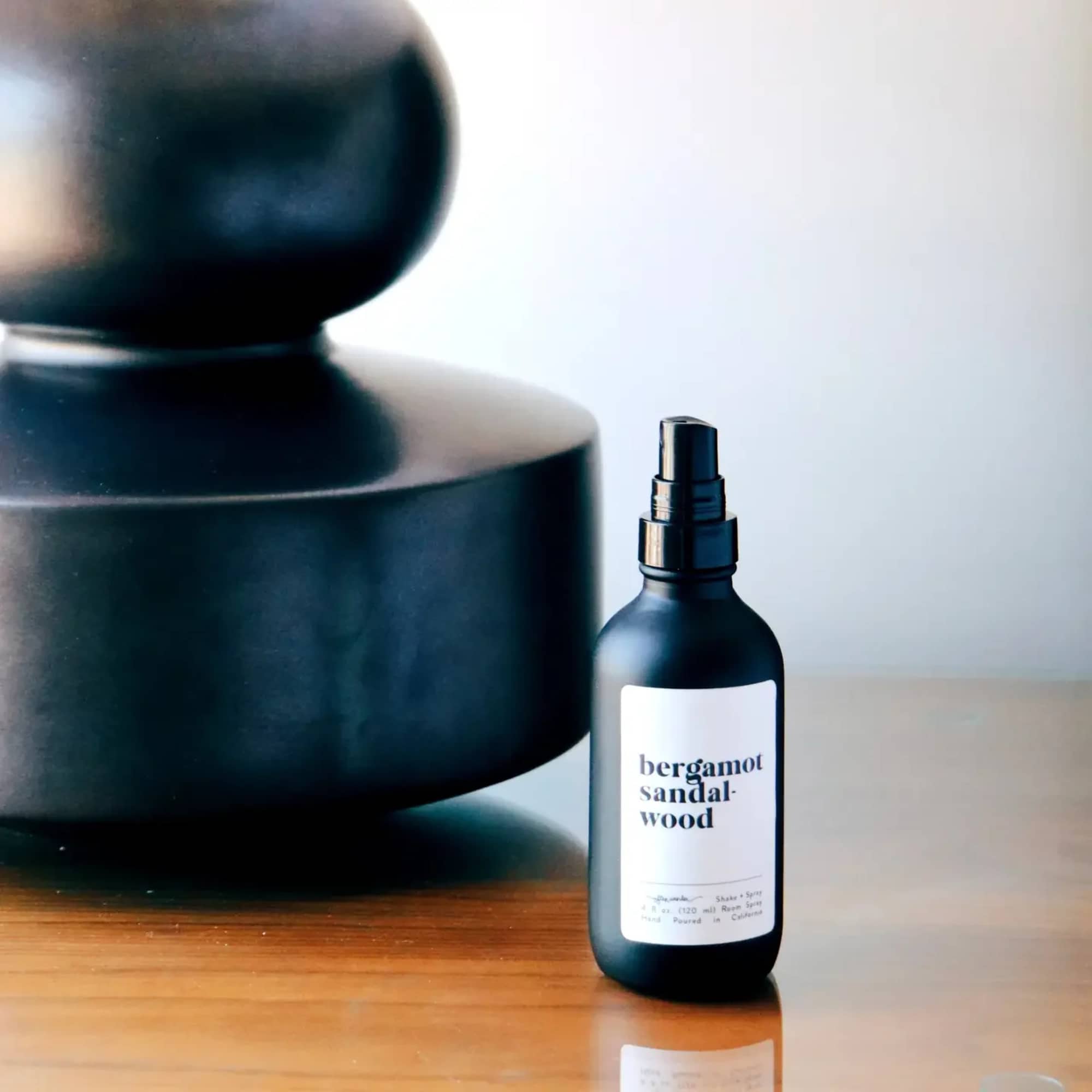 Matte black bergamot sandalwood spray bottle on a wood surface beside a large black decorative object
