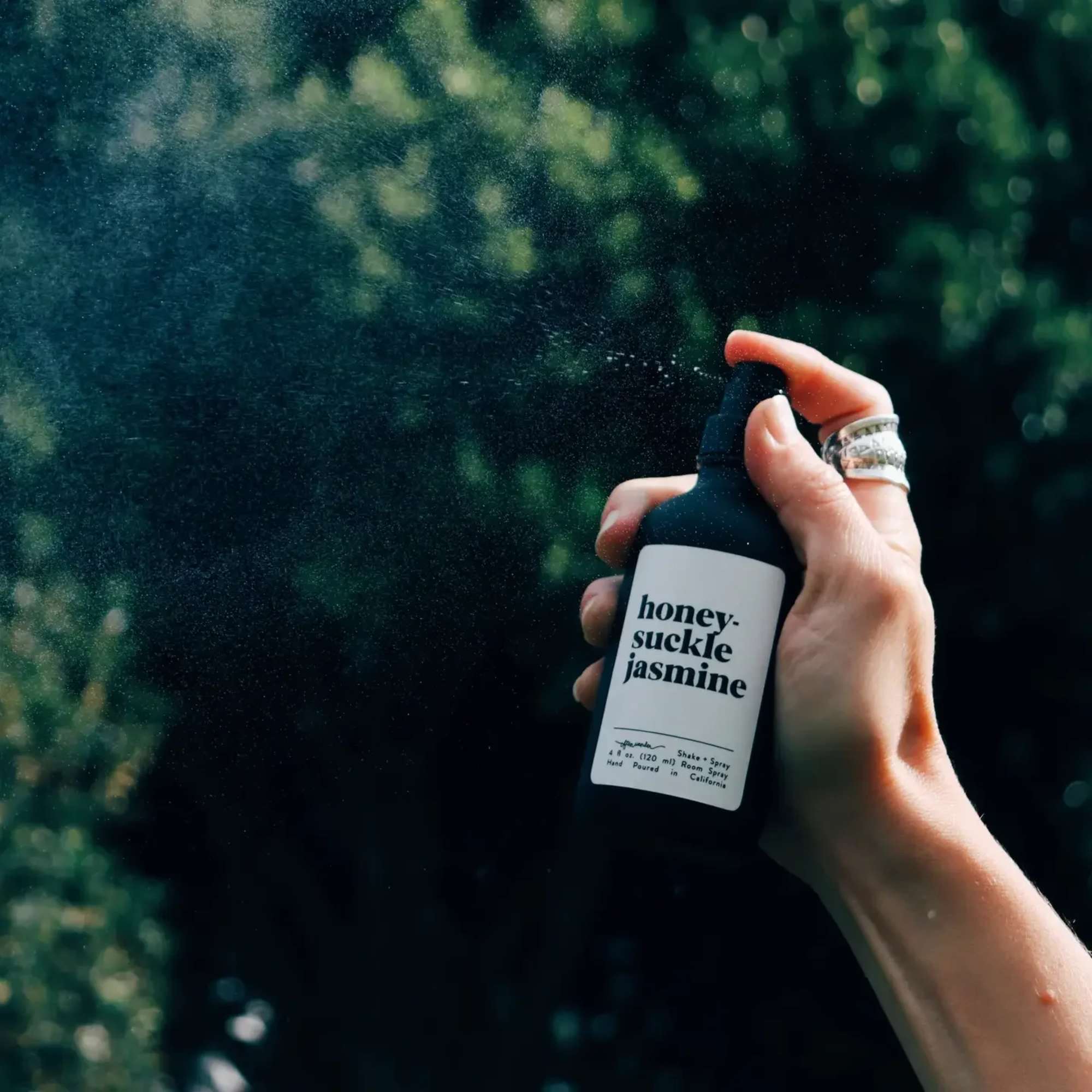 Hand spraying a black honeysuckle jasmine mist bottle against a blurred green background