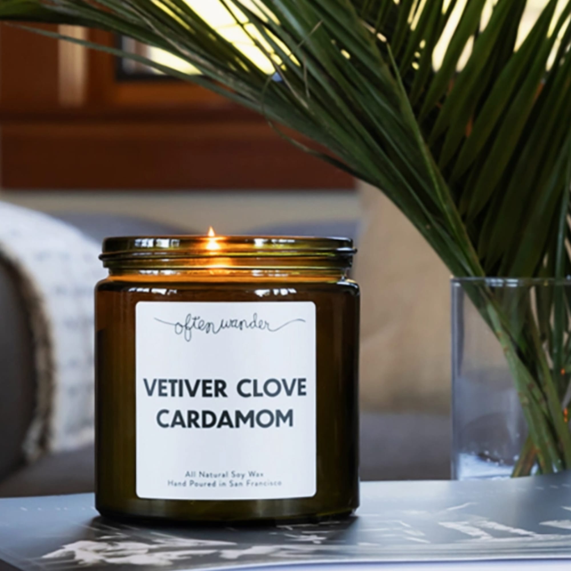Lit vetiver clove cardamom candle in an amber glass jar on a table with palm fronds in the background