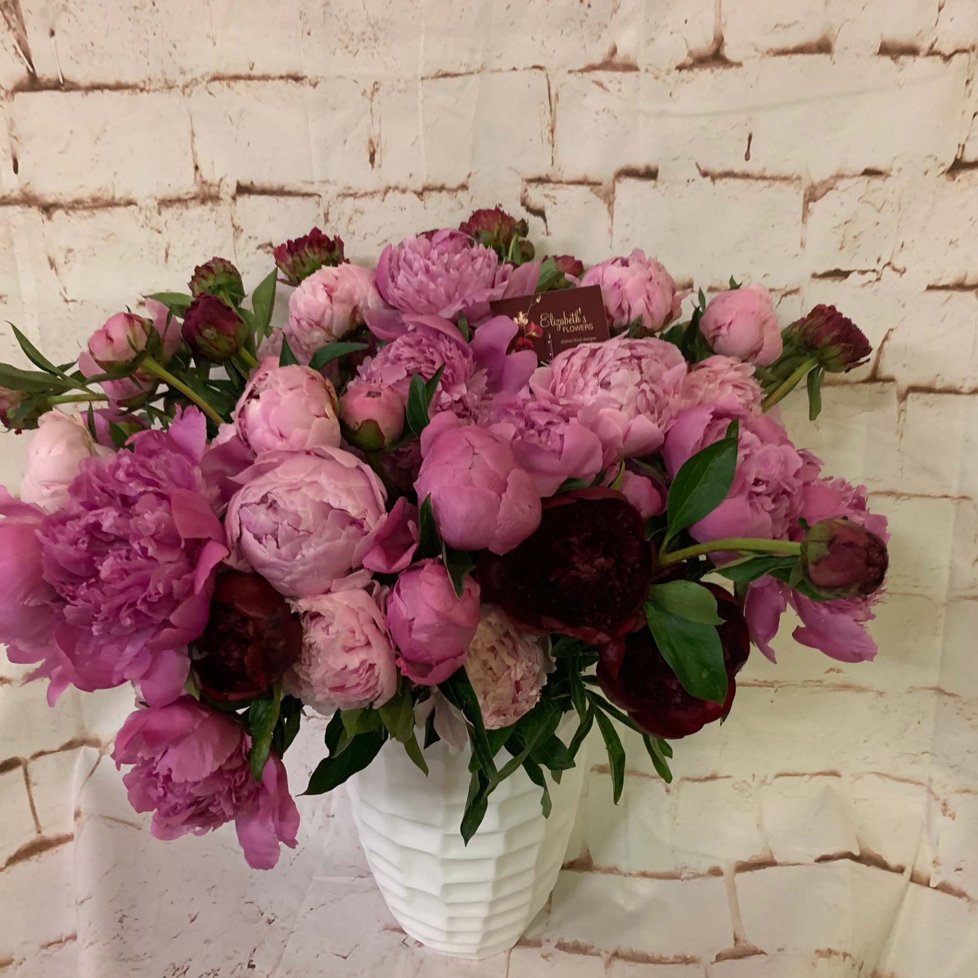 Lush arrangement of pink and burgundy peonies in a white textured vase against a light brick background.