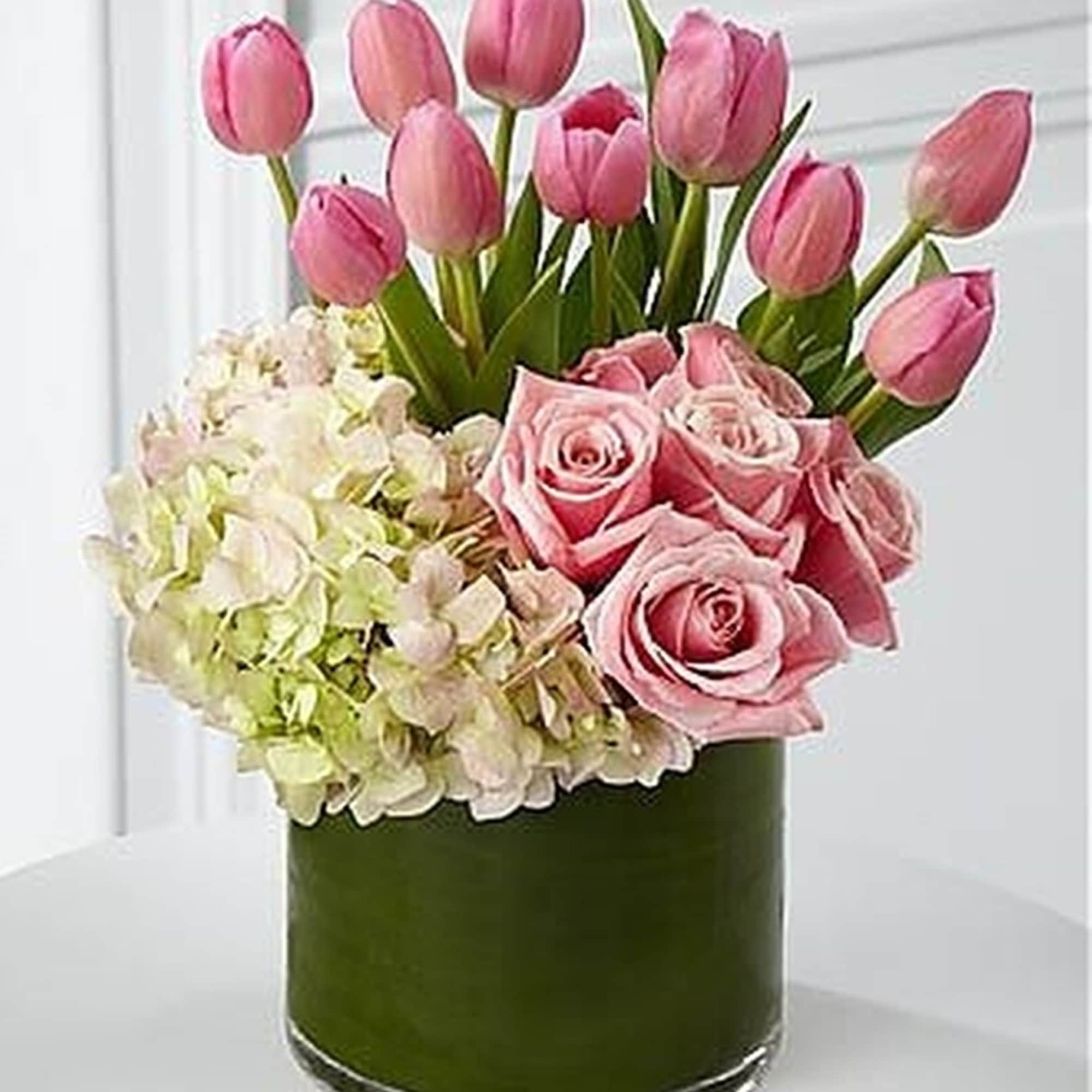 Pink tulips, roses, and pale hydrangeas arranged in a clear glass cylinder vase wrapped with green inside.