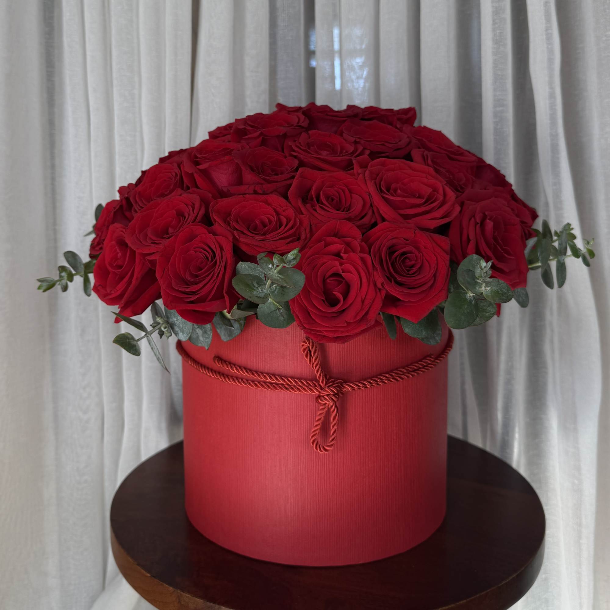 Red roses arranged in a round red box with eucalyptus accents