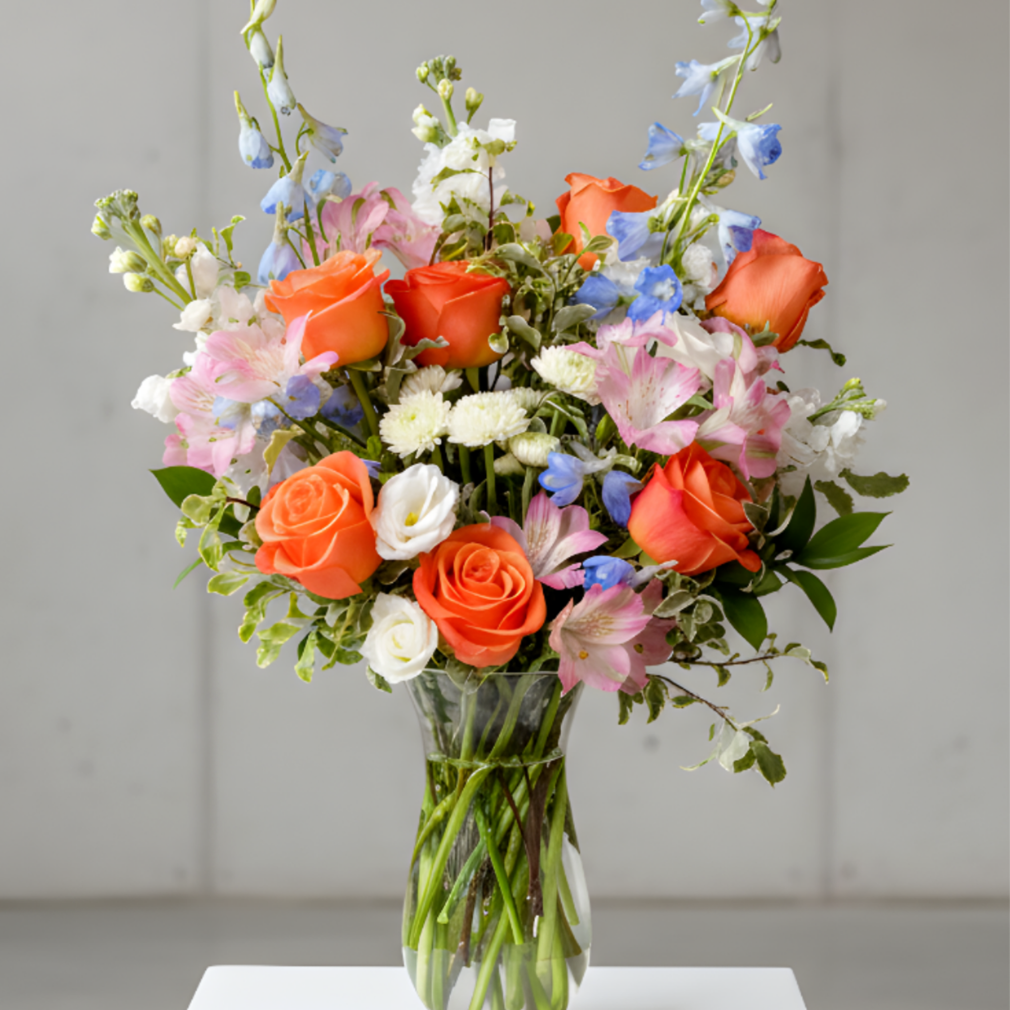 Tall mixed arrangement of orange roses, pink blooms, and blue delphinium in a clear glass vase