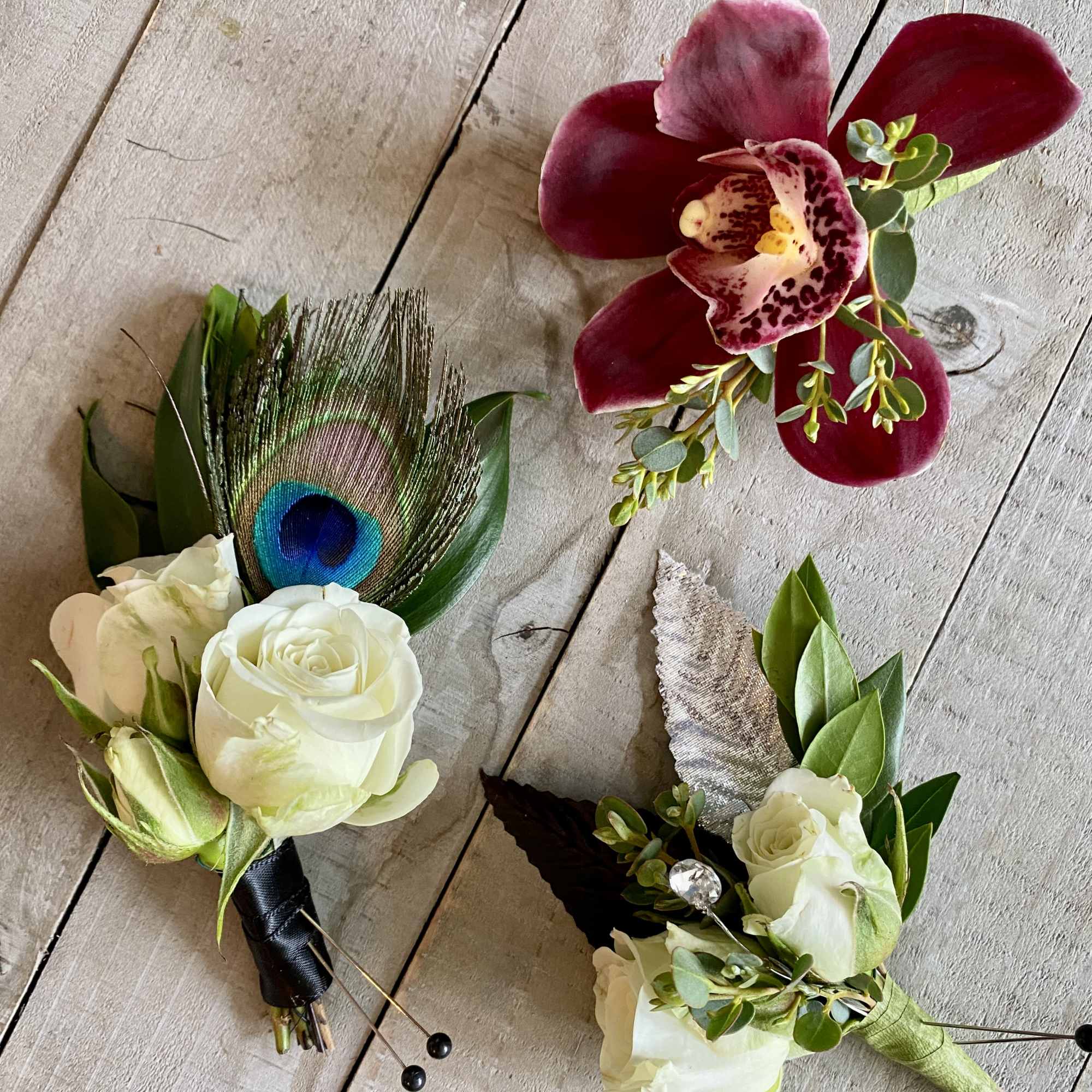 Three floral boutonnieres with white roses, burgundy orchids, and a peacock feather.