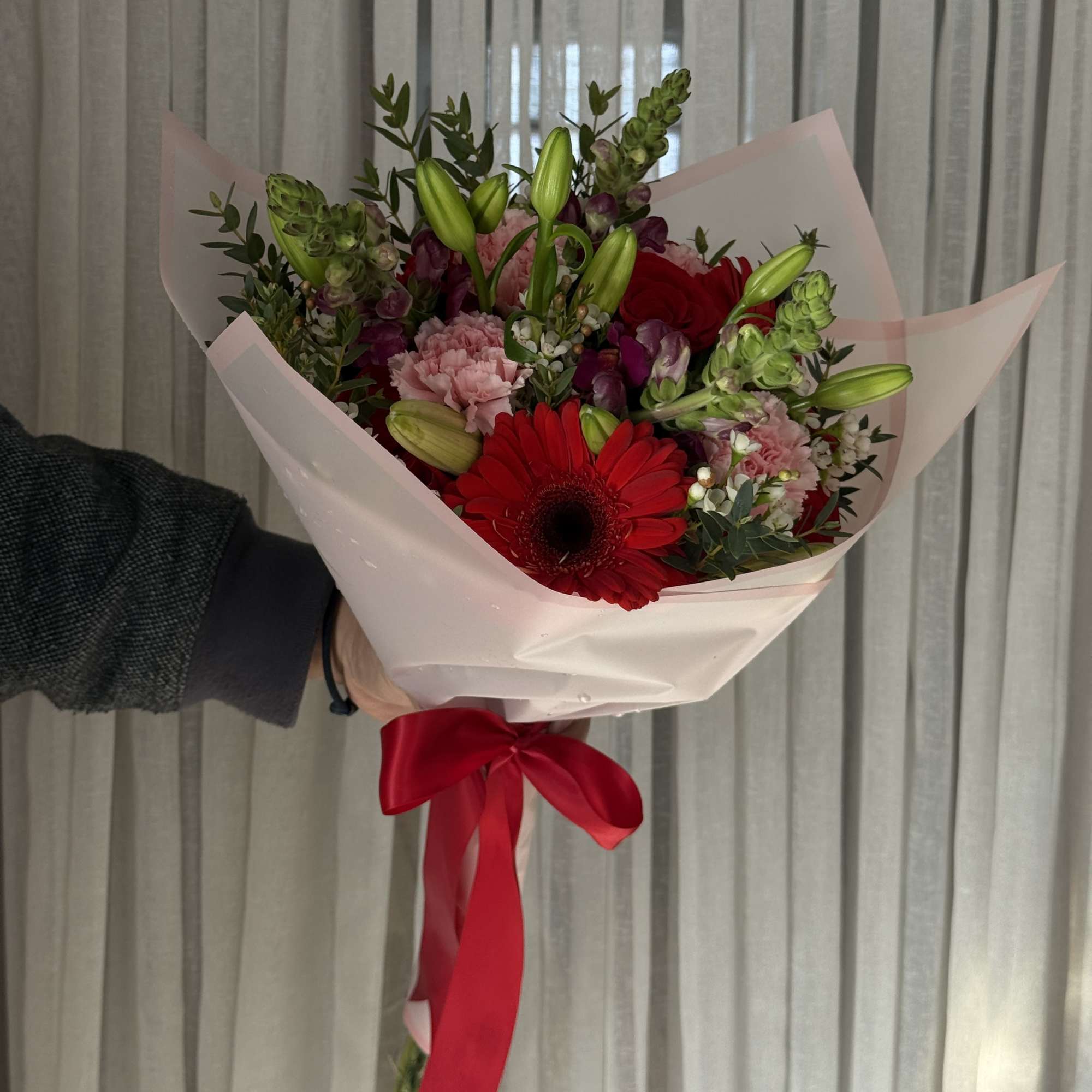 Handheld bouquet of red gerbera daisies, pink carnations, and lily buds wrapped in pale pink paper