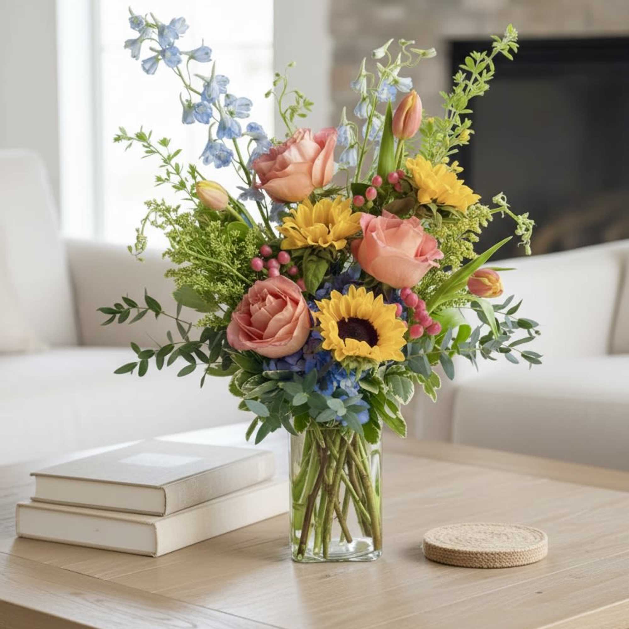 A clear glass vase arrangement with bright colors! Sunflowers, blue hydrangeas, orange/coral