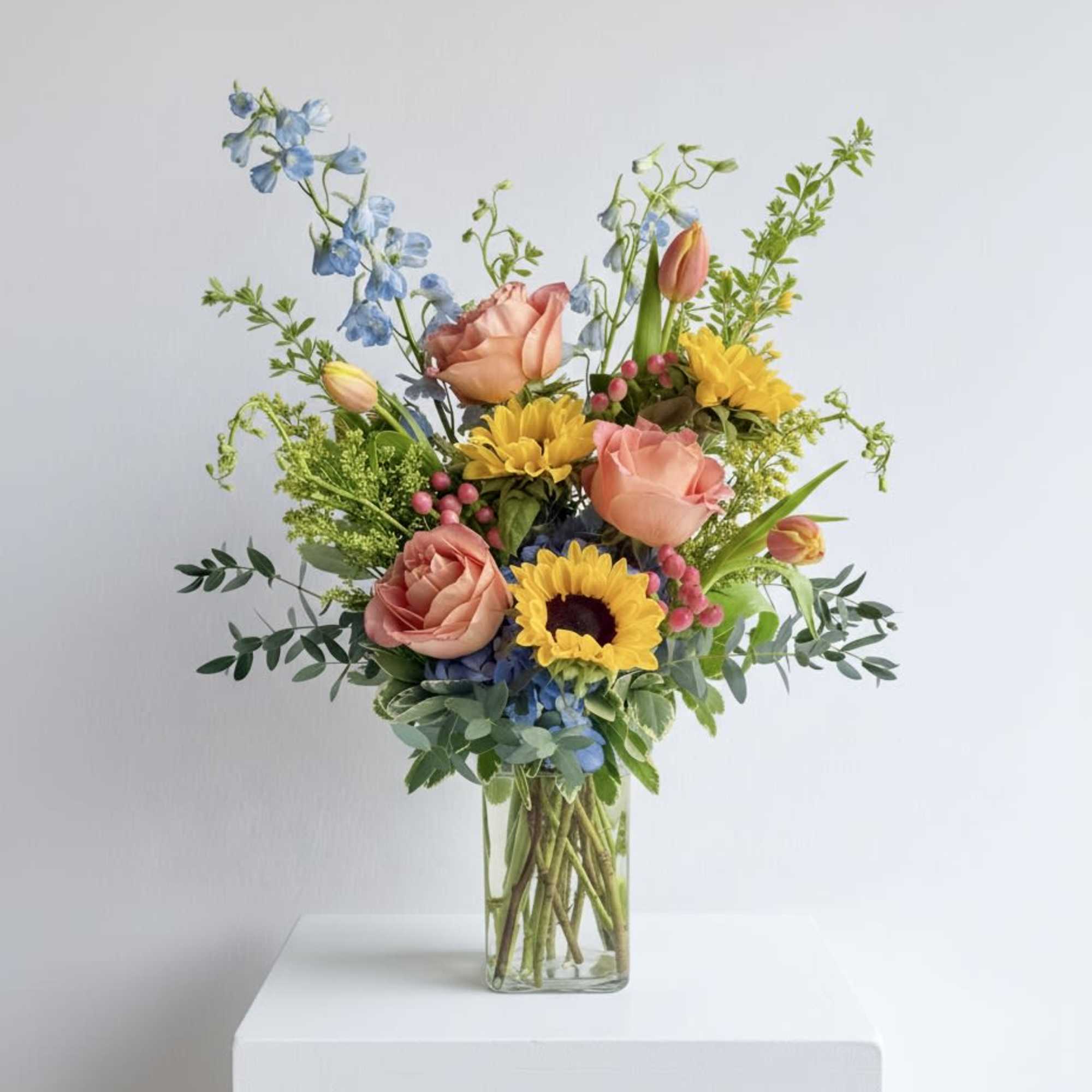 Tall mixed bouquet with yellow sunflowers, peach roses, blue blooms and tulips in a clear glass vase.