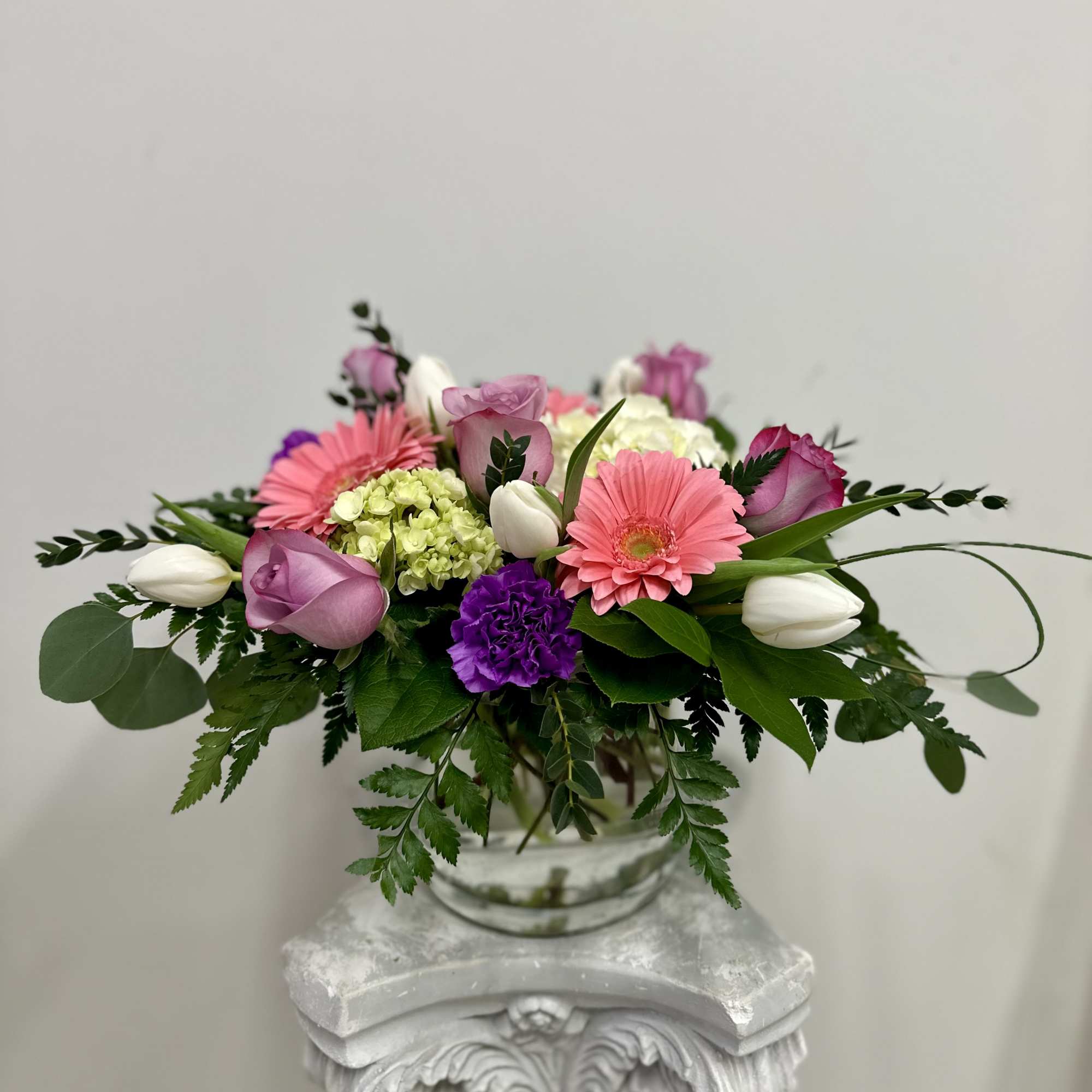 Low glass vase arrangement with pink gerbera daisies, purple roses, white tulips, and green hydrangea