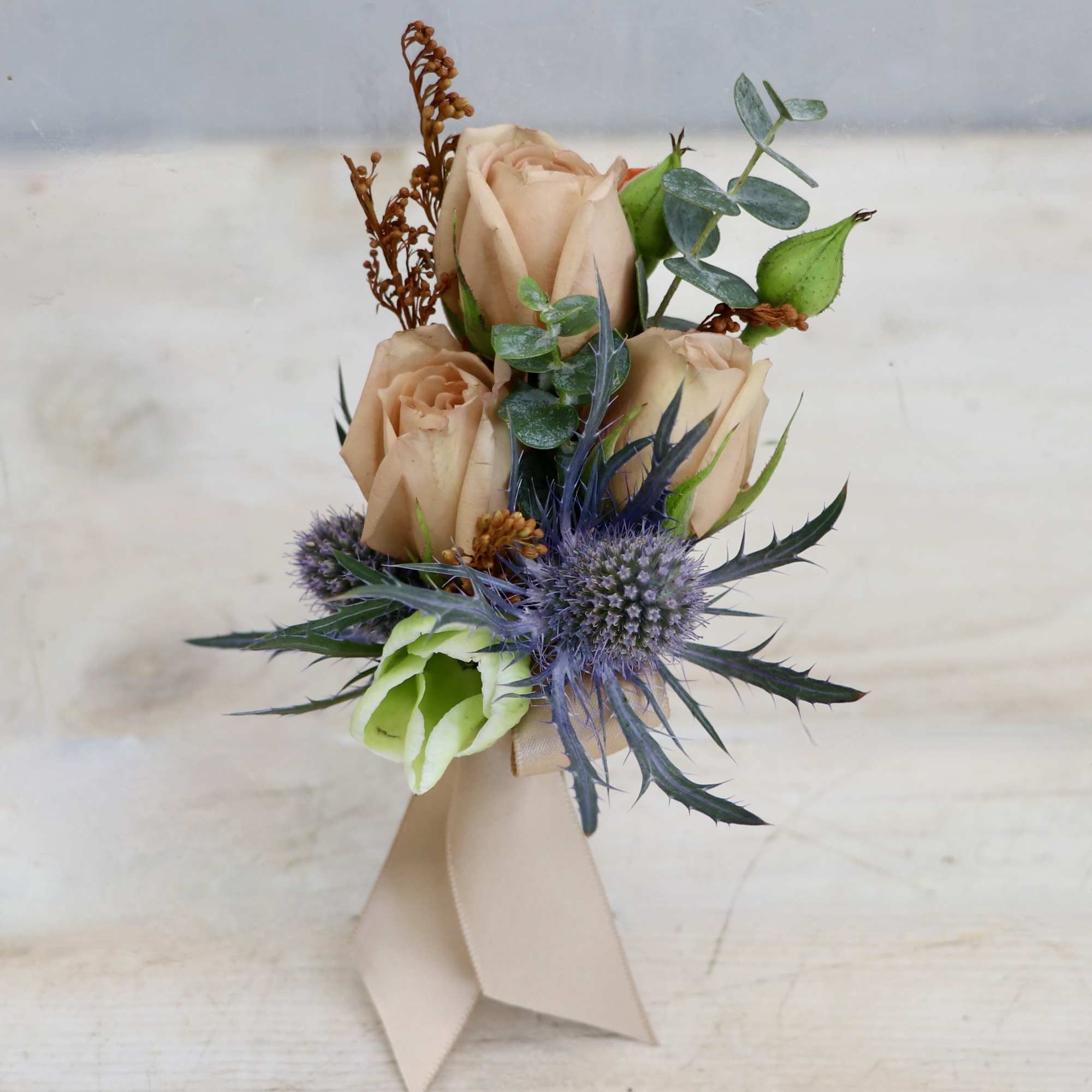 Peach rose boutonniere with blue thistle accents and beige ribbon wrap