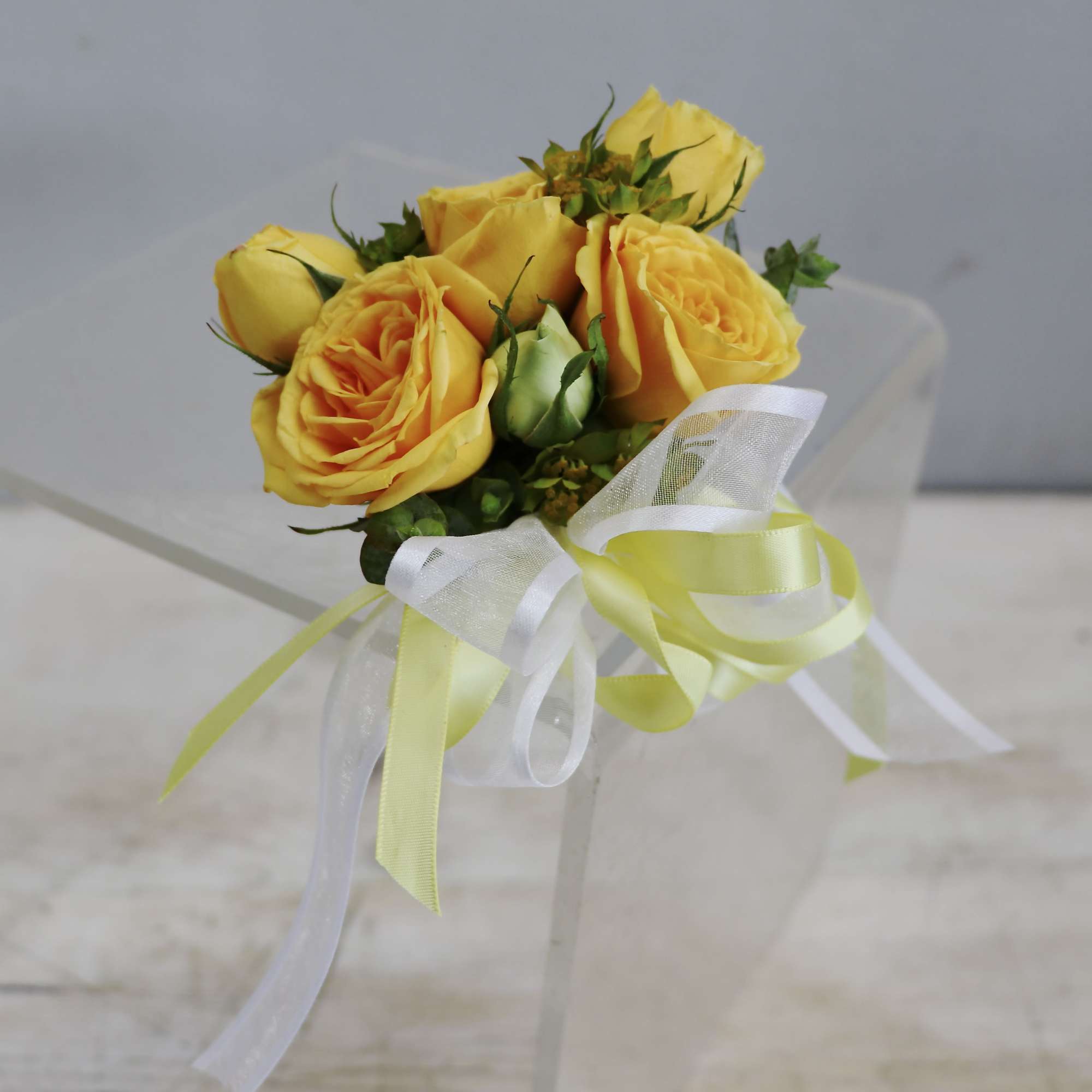 Yellow rose corsage with buds and white and yellow ribbon bow on a clear surface