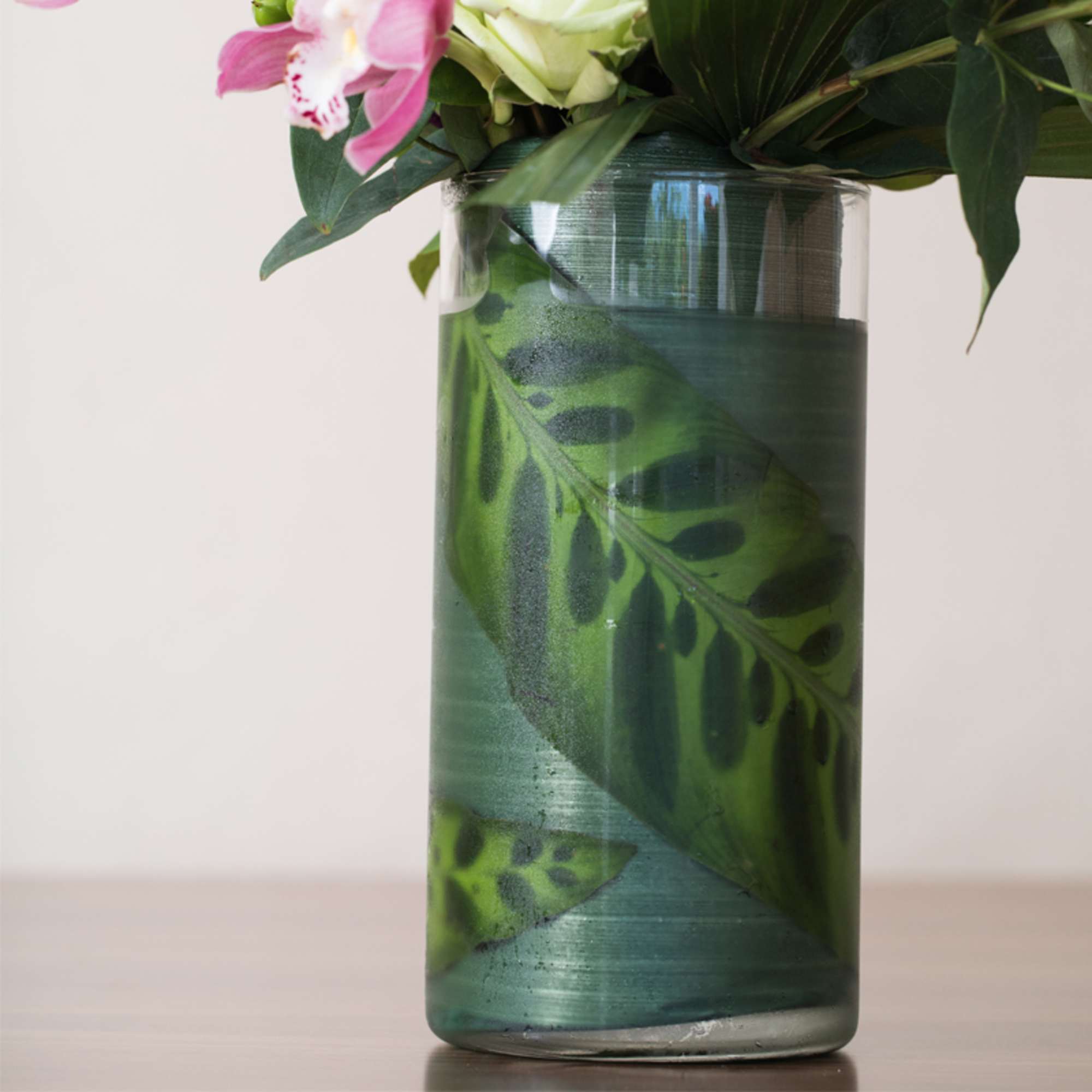 Tall clear glass vase with large patterned leaves inside and pink and white flowers at the top.