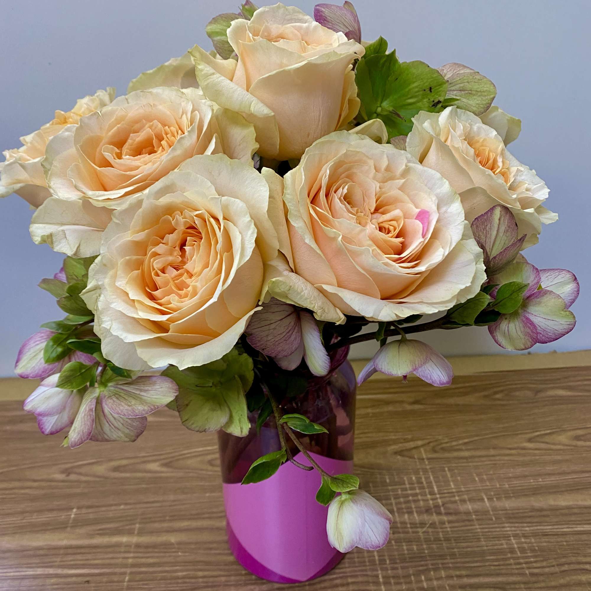 Peach roses arranged in a pink vase with pale green and purple blooms