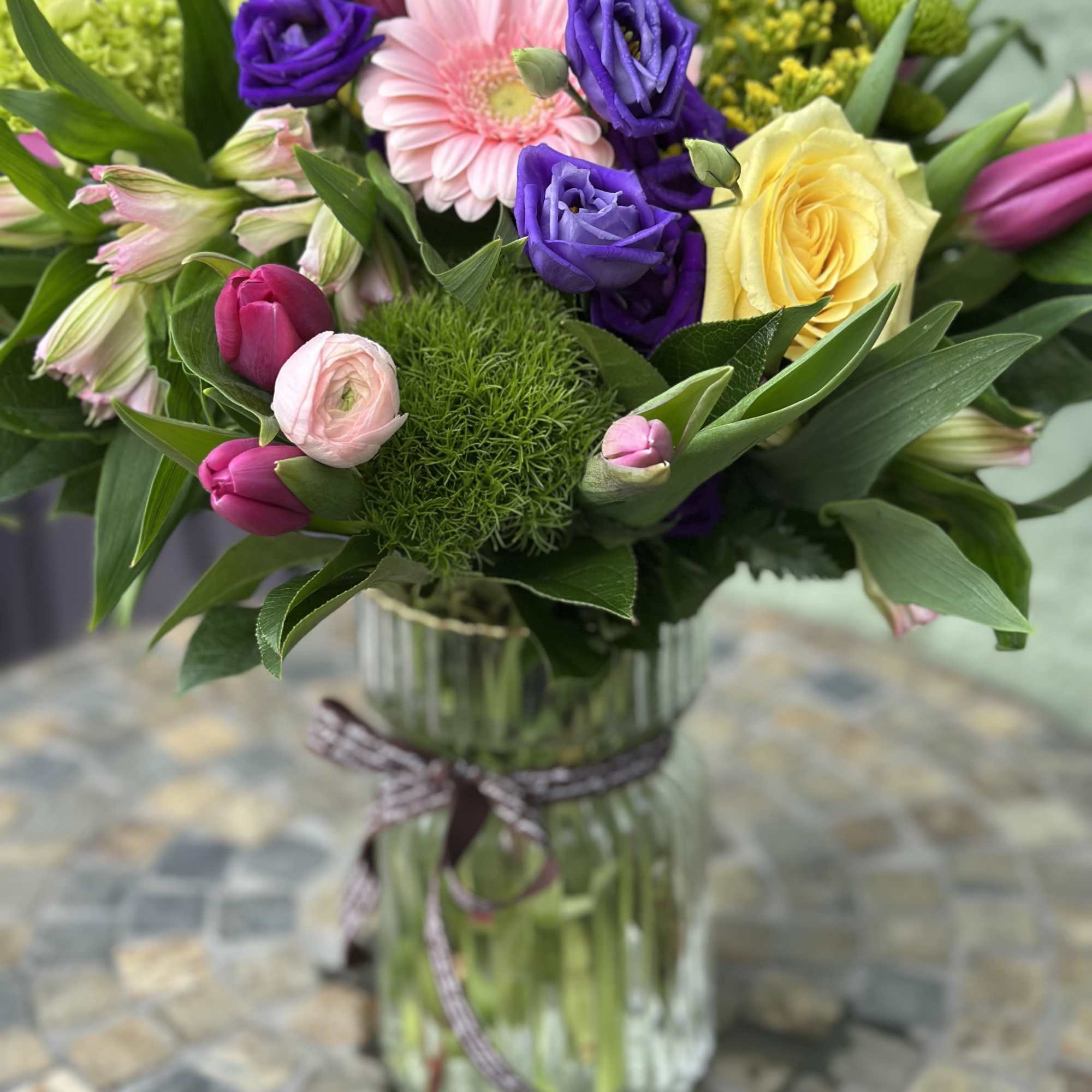 Mixed bouquet of pink, purple, yellow, and green flowers in a clear glass vase with a ribbon.
