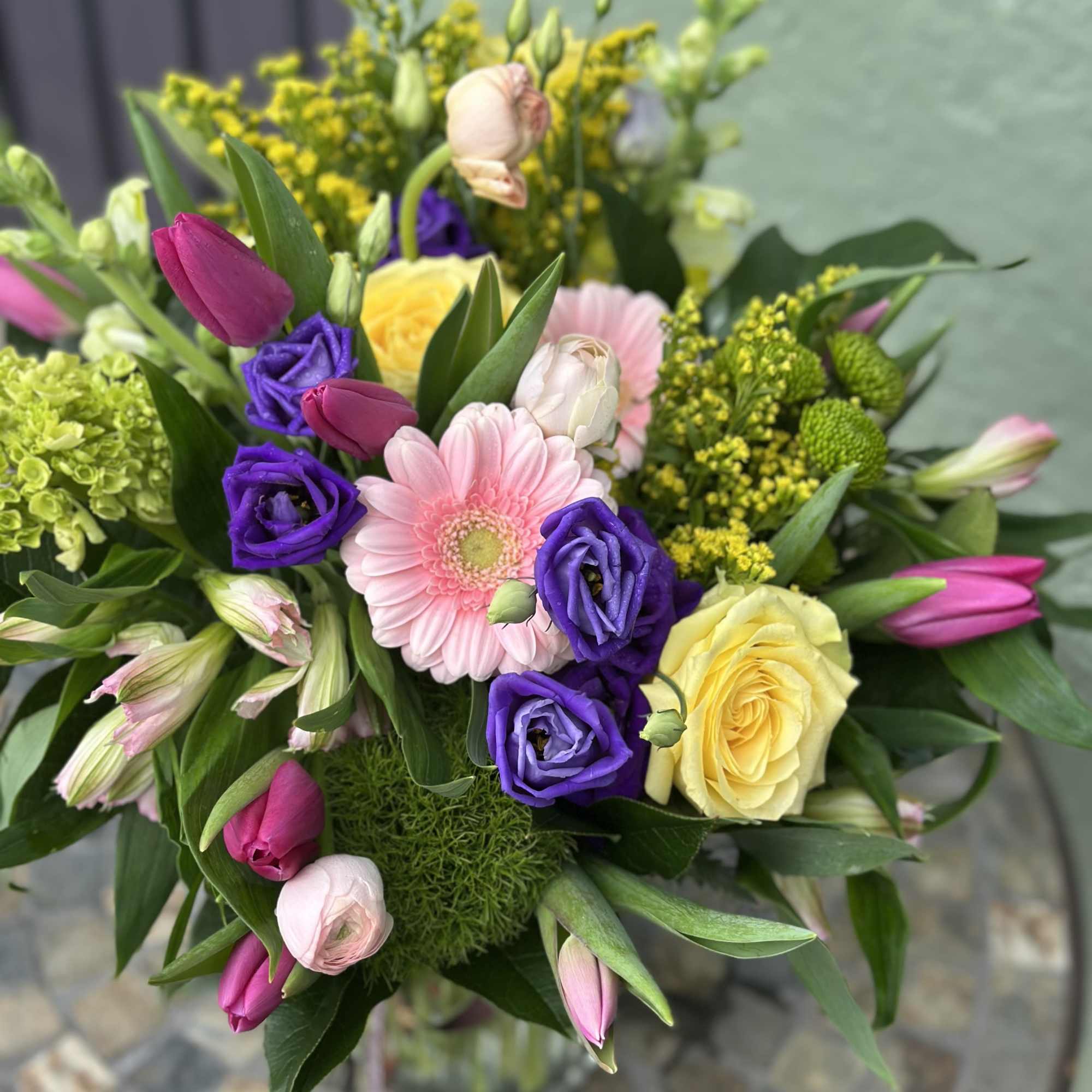 Bright mixed bouquet with pink tulips, purple blooms, yellow roses, and a pink gerbera in a clear glass vase.