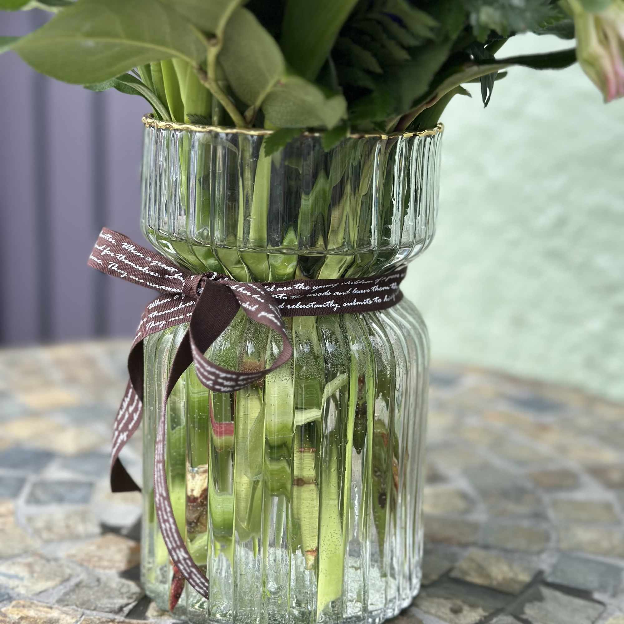 Clear ribbed glass vase with flower stems and a brown ribbon on a mosaic table